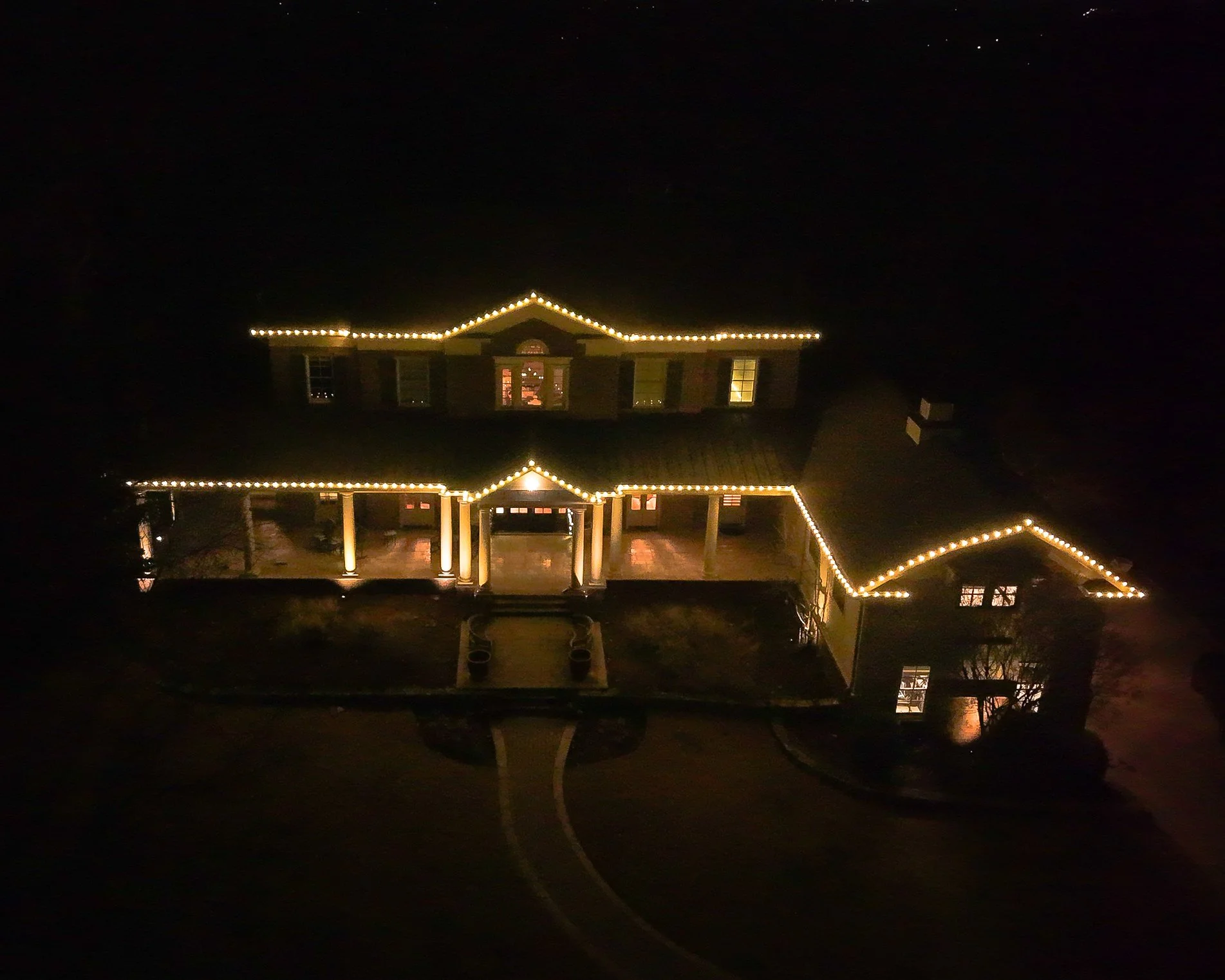 A large house decorated with string lights outlining the roofline, windows, and porch, illuminated at night, with a curved driveway leading up to it.