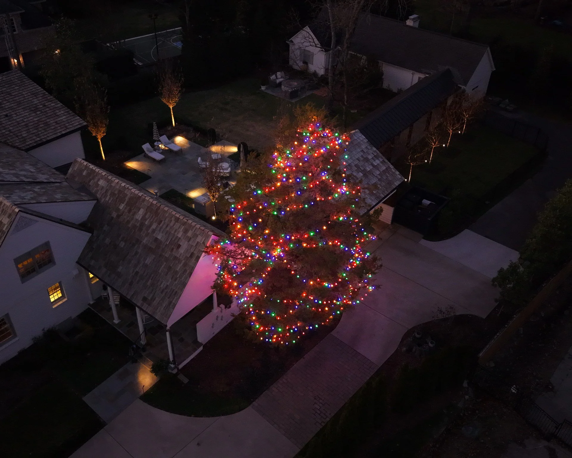 An aerial view of a neighborhood at night with a large tree decorated with multicolored Christmas lights in a driveway, surrounded by houses, trees, and a backyard with outdoor seating.