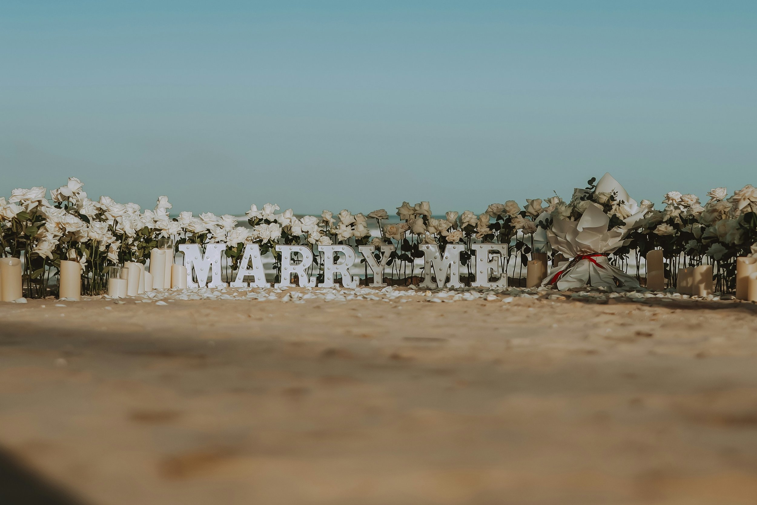 Beach wedding setup with white flowers, candles, and a large white sign that reads 'MARRY ME' on the sand.