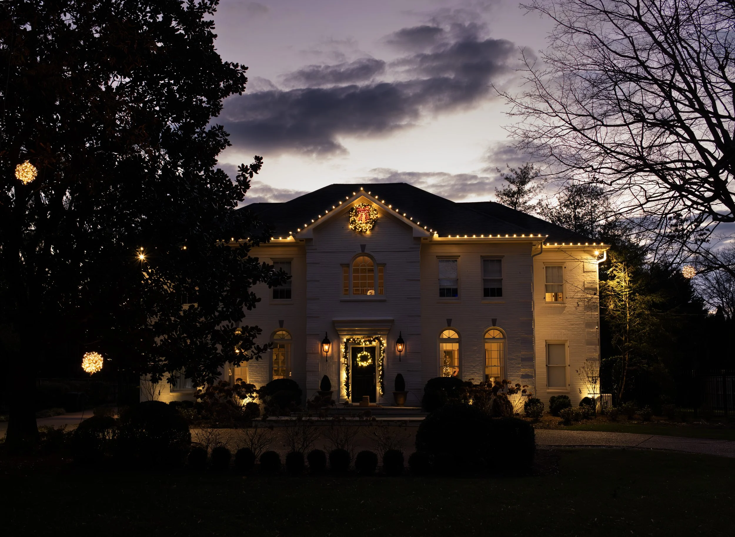 A large white house decorated with Christmas lights and wreaths, illuminated in the evening with a dark sky, trees, and clouds overhead.