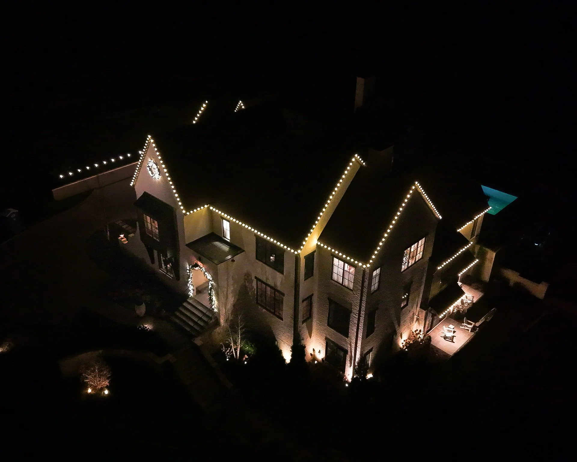 An illuminated house at night decorated with Christmas lights outlining the roof and windows, with a porch and outdoor seating area.