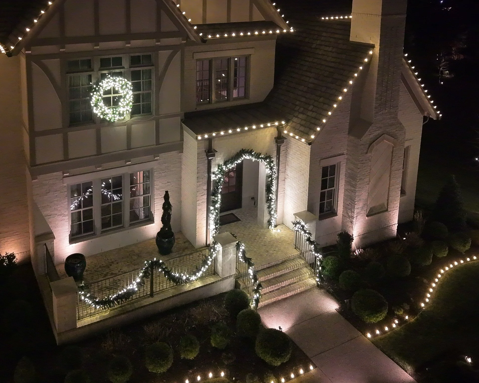 A large house decorated with Christmas lights and garlands. The house has a front porch with stairs, potted plants, and a gate, all adorned with white string lights and greenery. A wreath with lights hangs on a window above the porch, and a lit windo