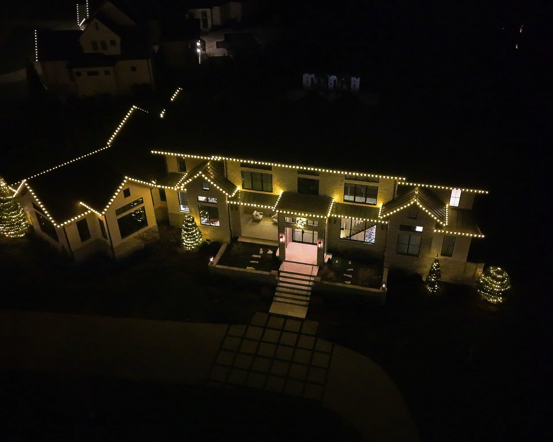 A large house decorated with string lights outlining the roof and windows, with illuminated Christmas trees outside, taken at night.