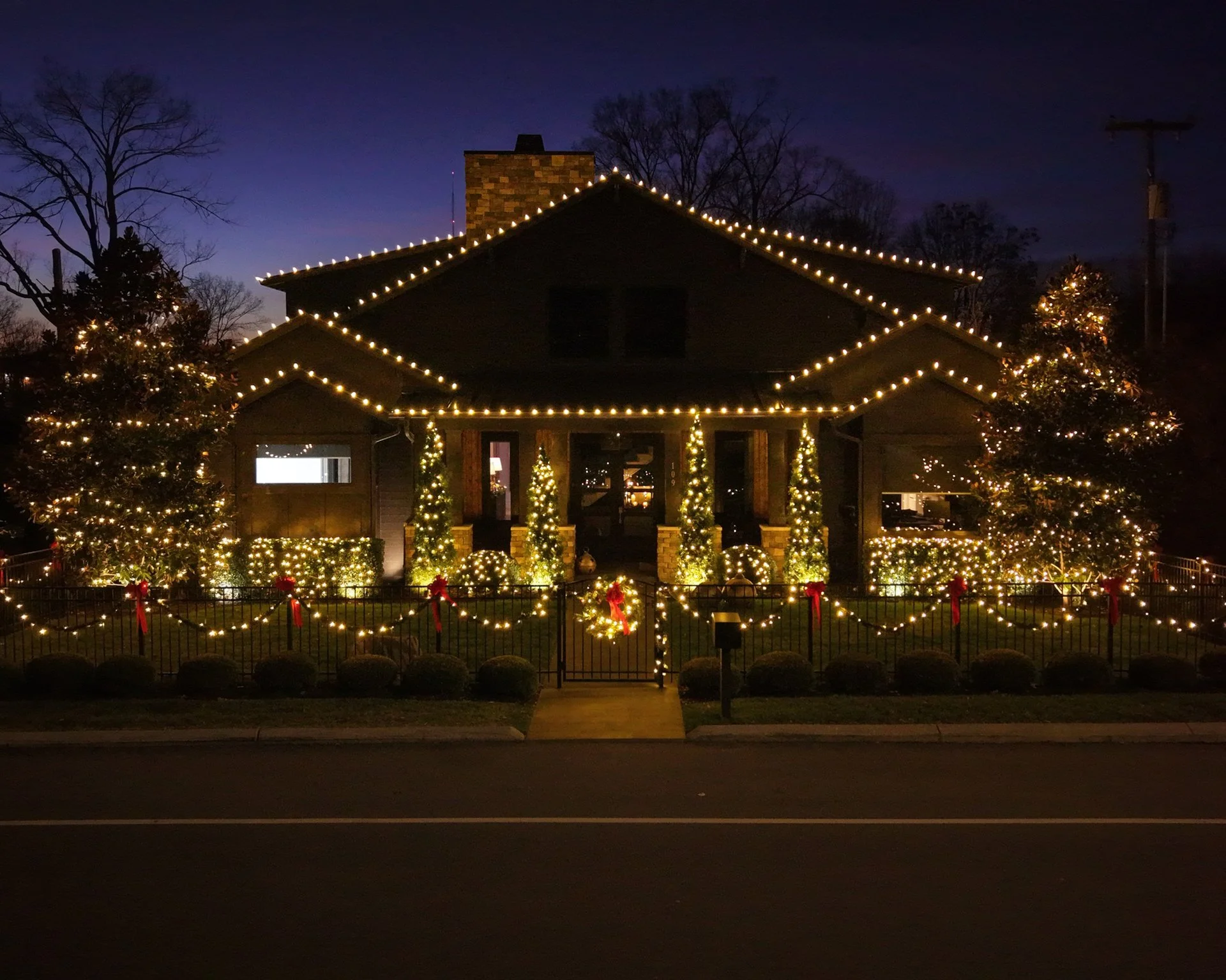 A house decorated for Christmas with string lights outlining the roof, trees, and porch, with additional lights wrapped around bushes and in the yard at night.