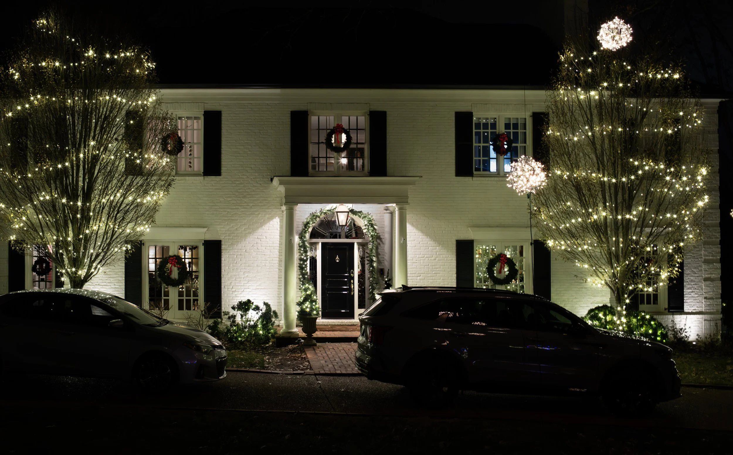 A white house decorated with Christmas wreaths, lights, and ornaments at night, with two black cars parked in front and illuminated trees on either side.