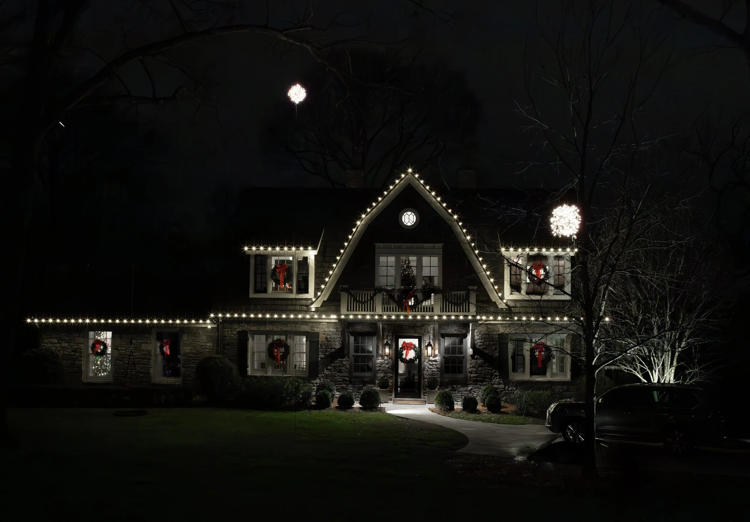 A house decorated for Christmas with white string lights outlining the roof and windows, red wreaths, and a Christmas tree visible through the window, illuminated at night with a full moon in the sky.
