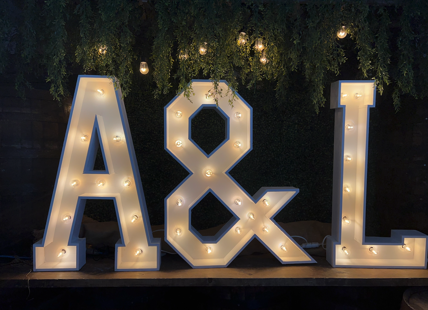 Large illuminated marquee letters spelling 'A&L' with hanging string lights and hanging greenery above.
