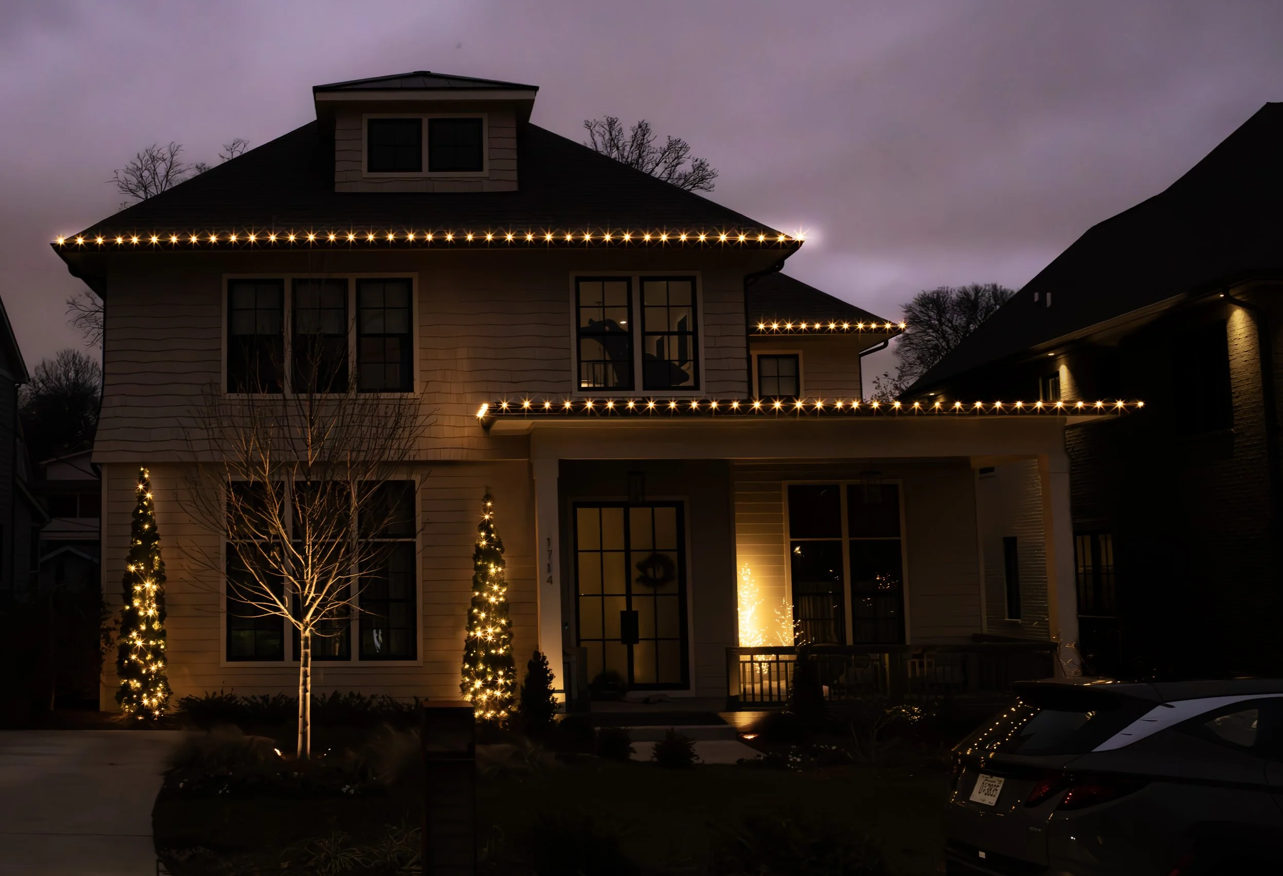 A two-story house decorated with Christmas lights at night. The house has string lights along the roofline, illuminated trees in the front yard, and a Christmas tree visible through a window.
