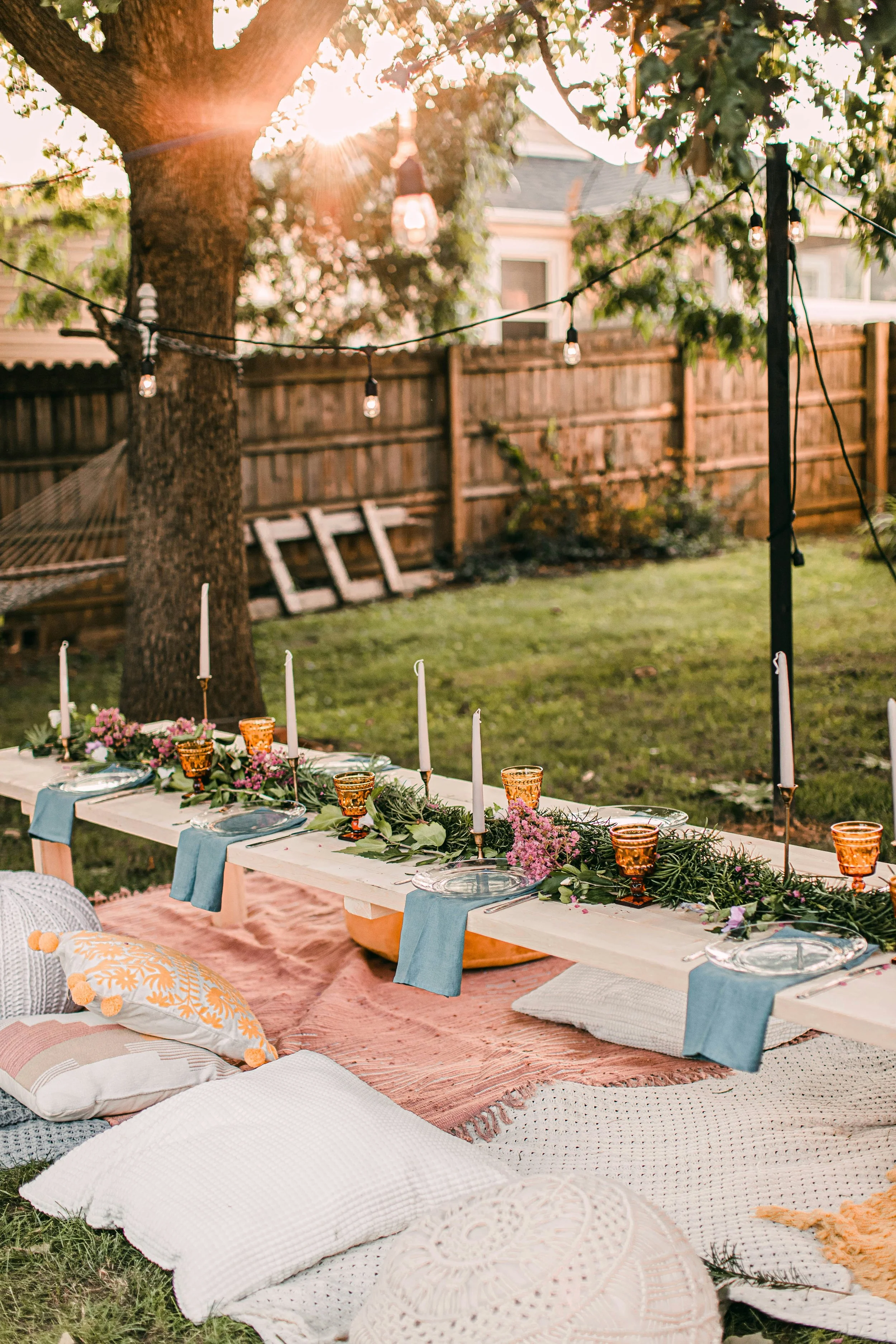 An outdoor dinner setup with a long table decorated with candles, flowers, and tableware, set in a backyard with a tree, string lights, and a wooden fence during sunset.