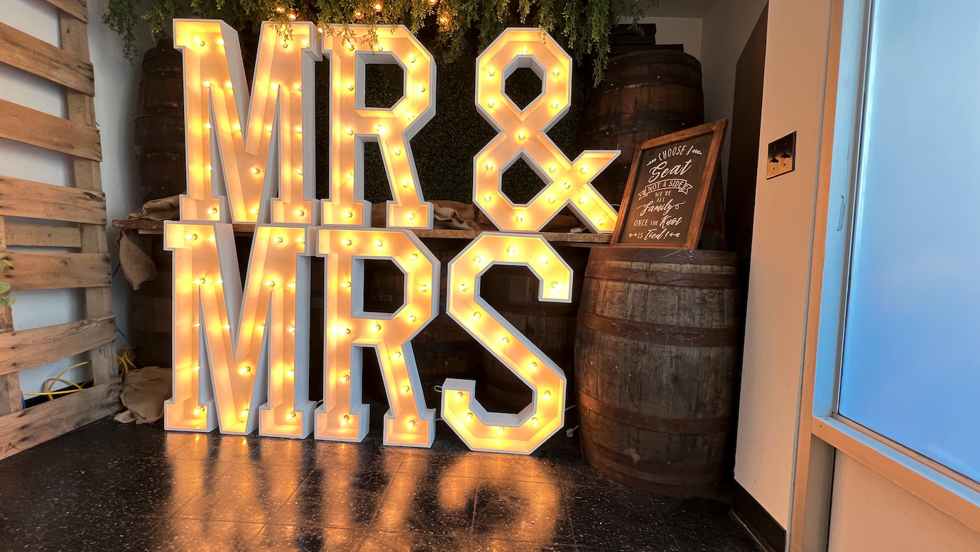 Large illuminated marquee letters spelling 'MR & MRS' on a shelf, with barrels and a framed sign beside them, in a rustic decor setting.