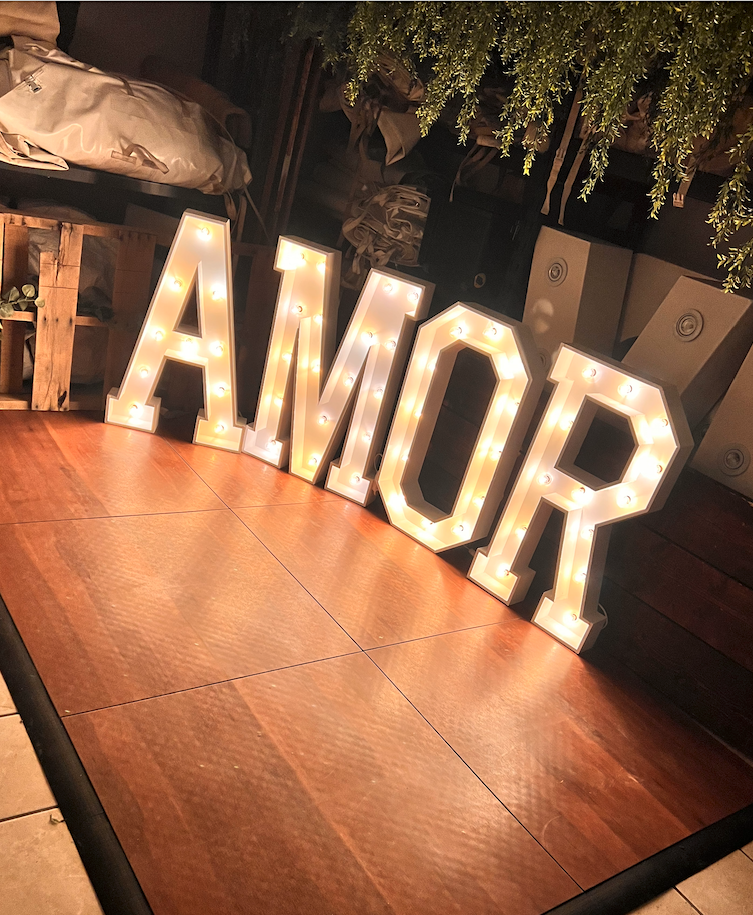 Illuminated marquee letters spelling 'AMOR' on a wooden floor in a cozy indoor setting.