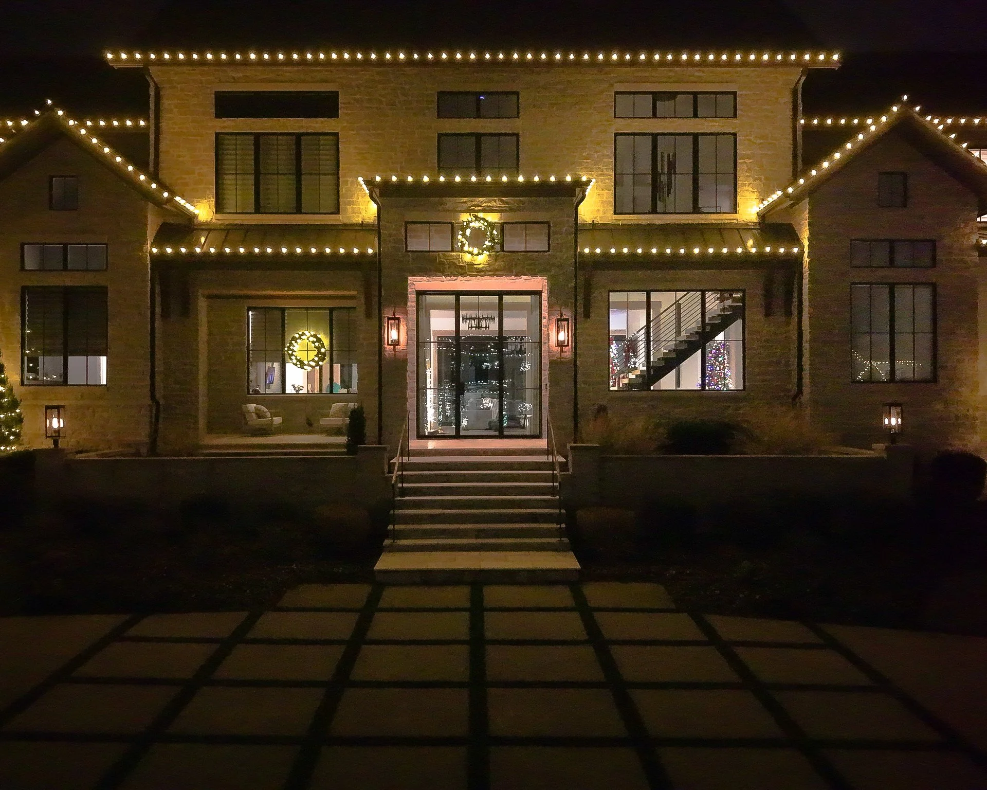 A large house decorated with white Christmas lights, wreaths, and a Christmas tree visible inside through the windows at night.