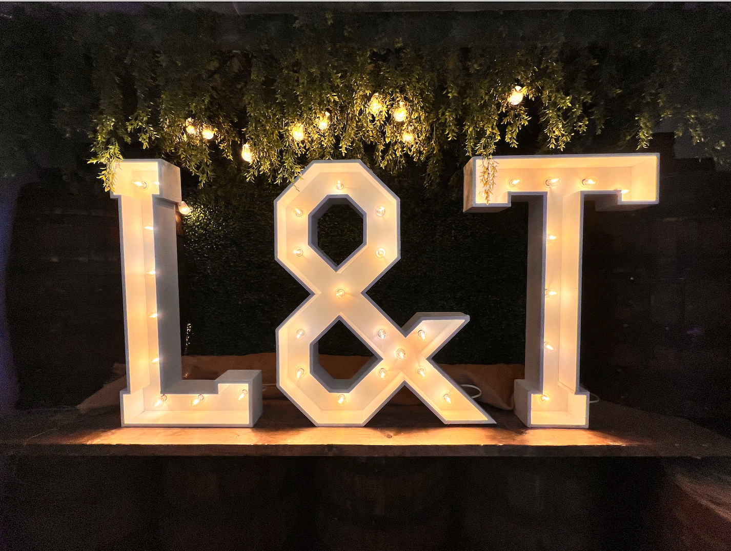  illuminated marquee letters spelling 'L & T' on a wooden surface with a dark background, greenery, and hanging string lights above.