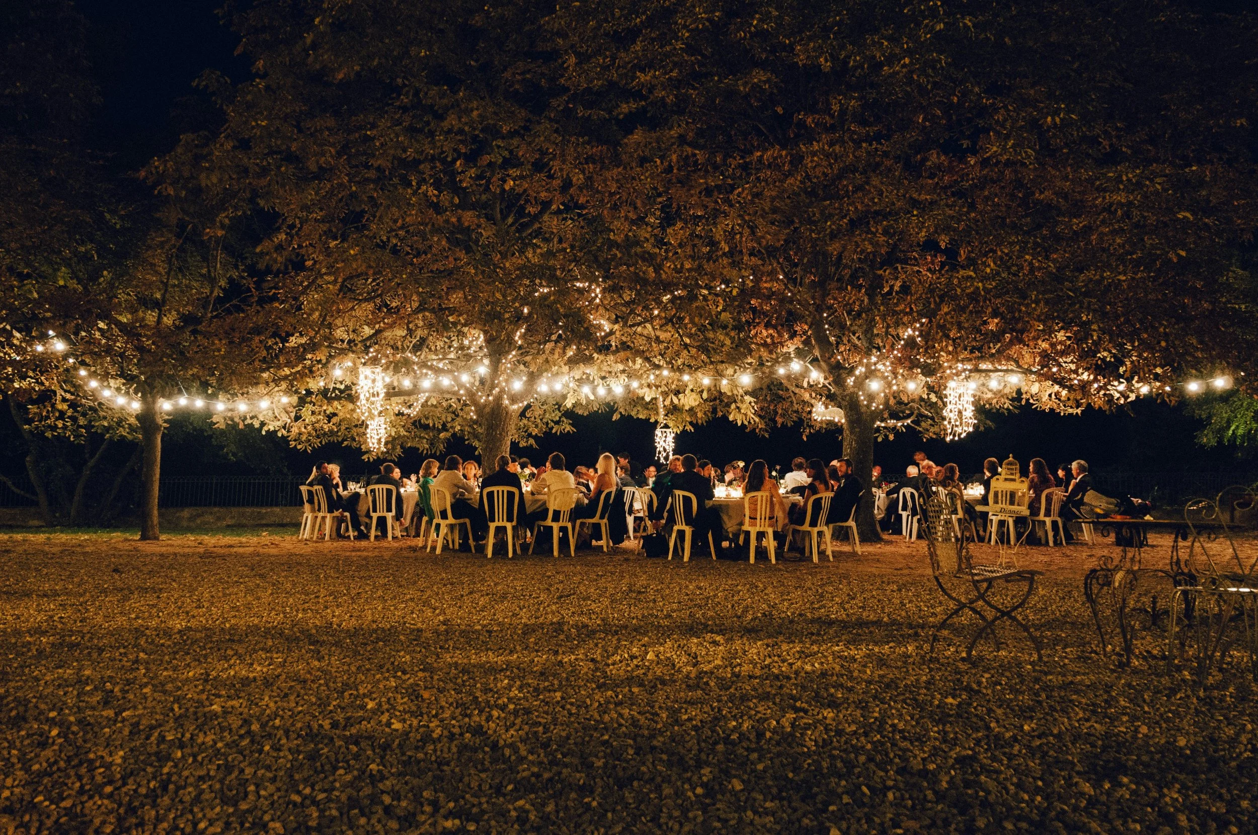 Outdoor nighttime dinner party under large trees decorated with string lights, with multiple tables and chairs, and guests enjoying the event.