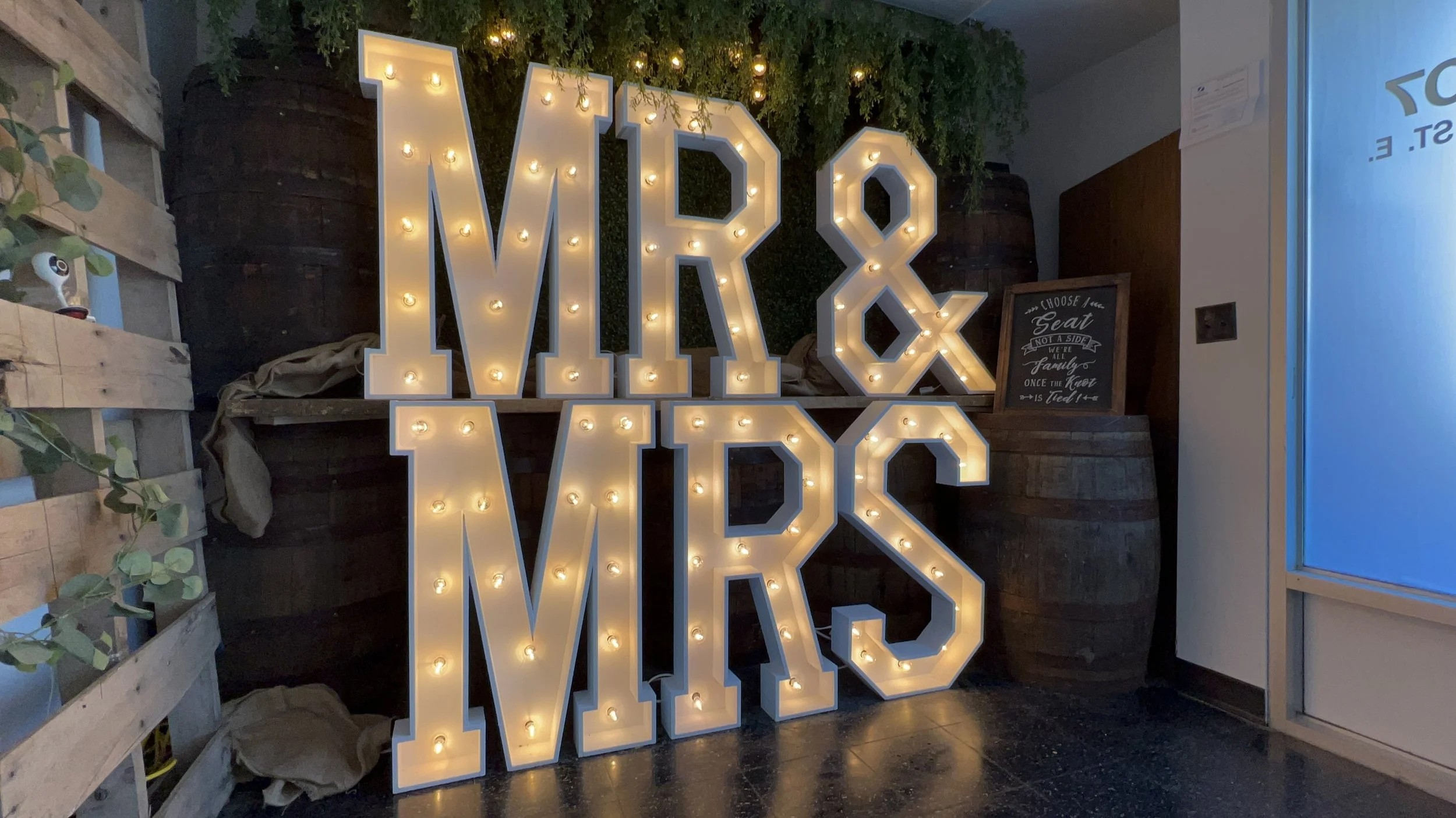 Large illuminated marquee sign reading 'MR & MRS' with decorative greenery and barrels in the background.
