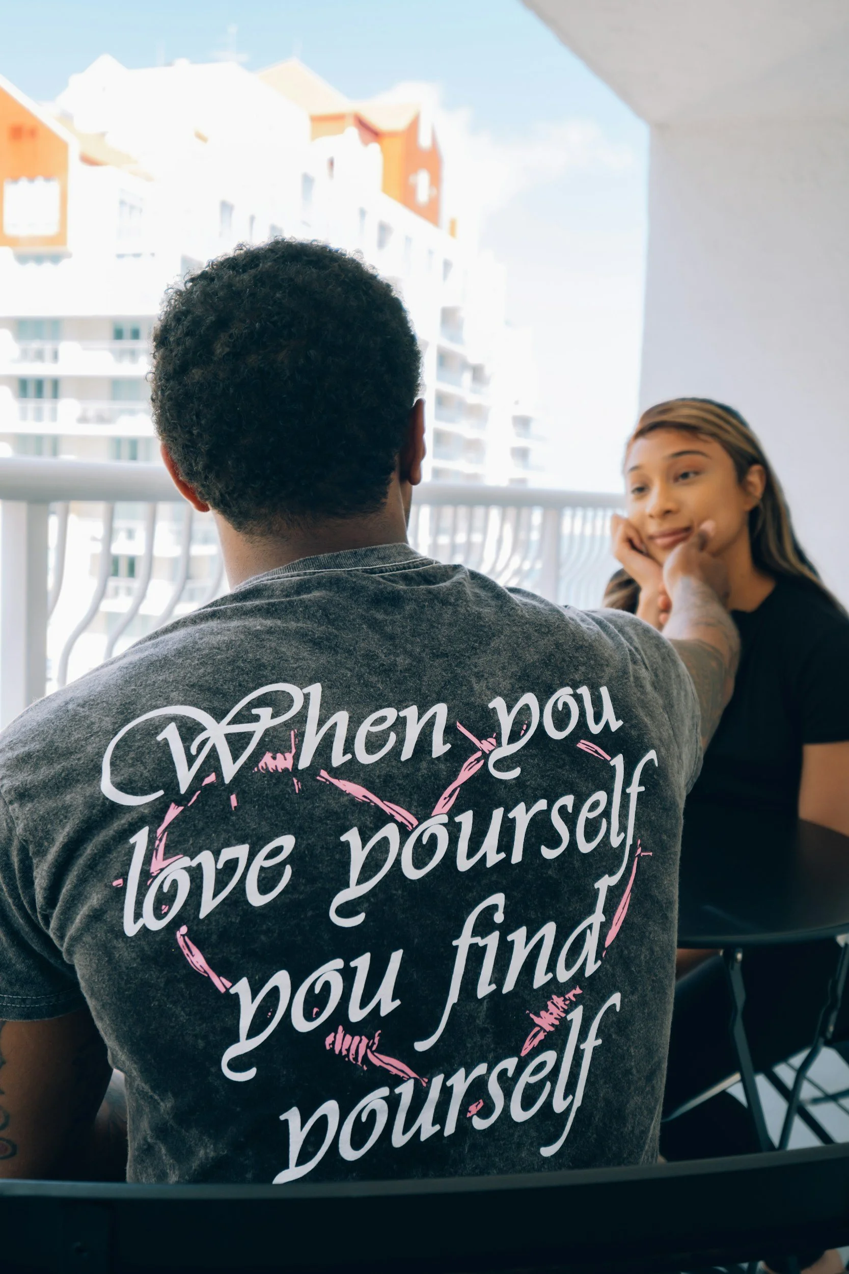 A man with curly hair wearing a gray T-shirt with a pink heart and the quote, 'When you love yourself, you find yourself,' is touching the face of a woman with long brown hair, sitting on a balcony with the city in the background.