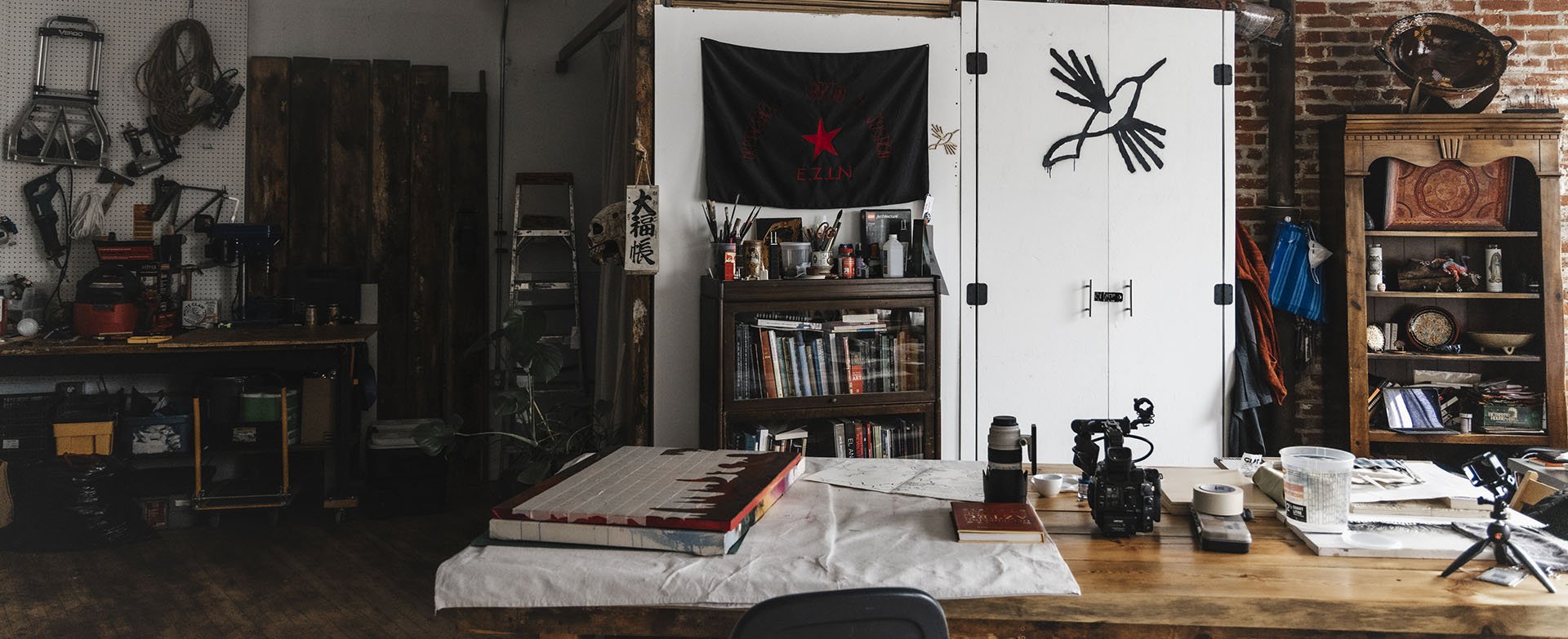A studio workspace  with equipment, tools, books, and decorative items, including a EZLN flag on the wall, a wooden cabinet, and a white door with hand-painted black Colibri logo.