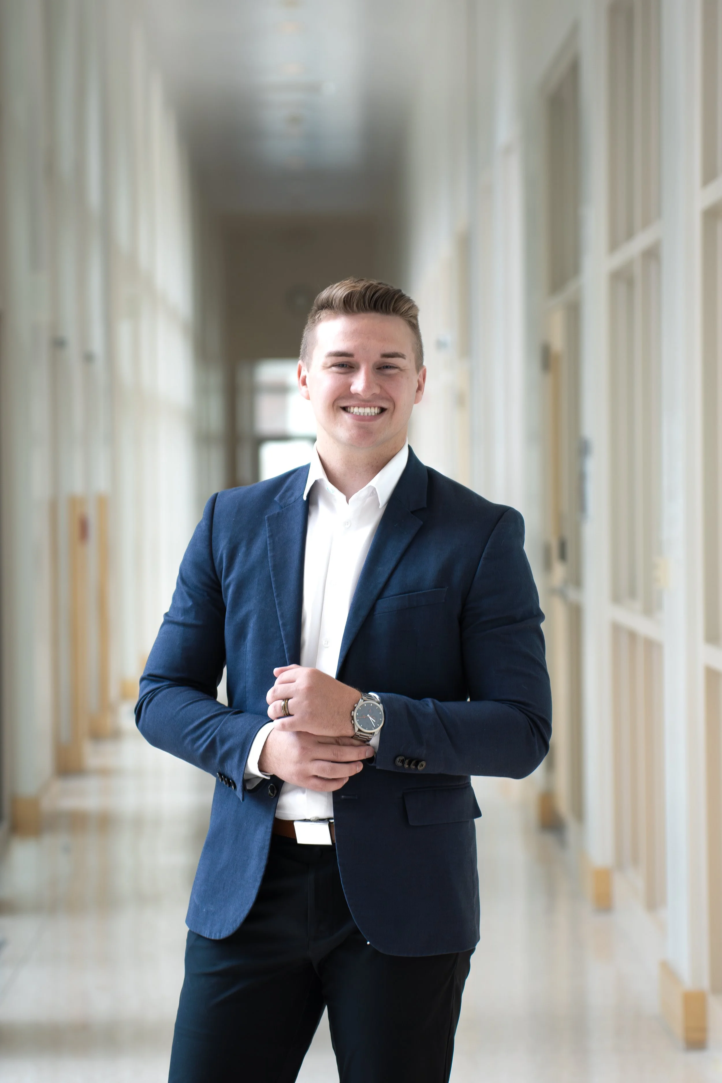 A young man in a navy blue suit and white shirt standing in a bright, corridor with glass display cases and large windows, smiling with arms crossed.
