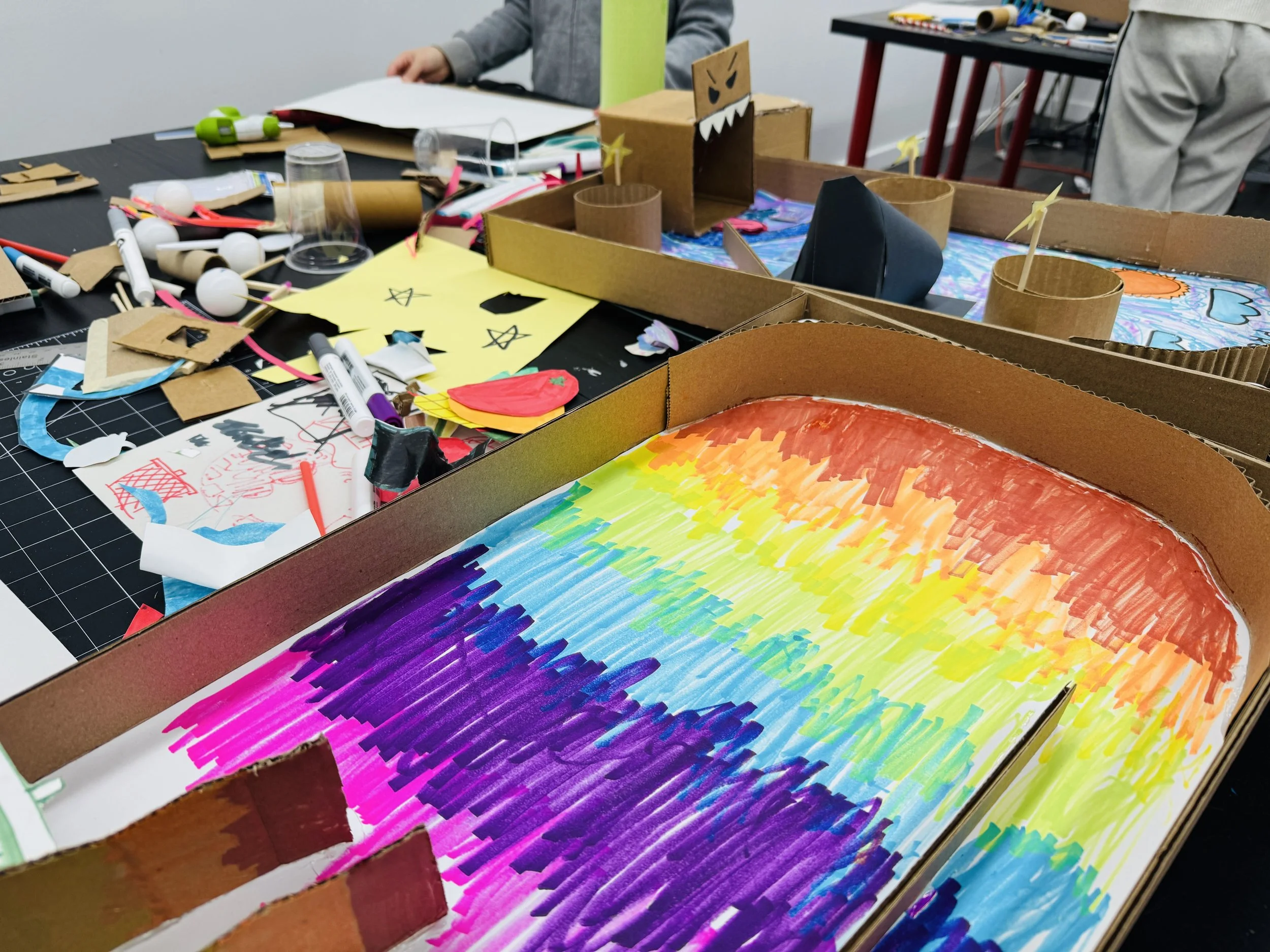 A tabletop covered with art supplies, colorful drawings, a cardboard maze, and a painted rainbow scene, indicative of a creative project or workshop.