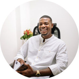 A young man with a bright smile sitting in an office chair, holding a smartphone and a calculator, with a flower arrangement in the background.