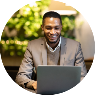 A man in a gray blazer and sweater smiling while working on a laptop outdoors with green foliage in the background.