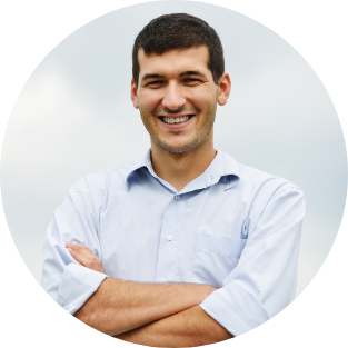 A smiling man with dark hair and a light blue shirt standing outdoors against a cloudy sky.
