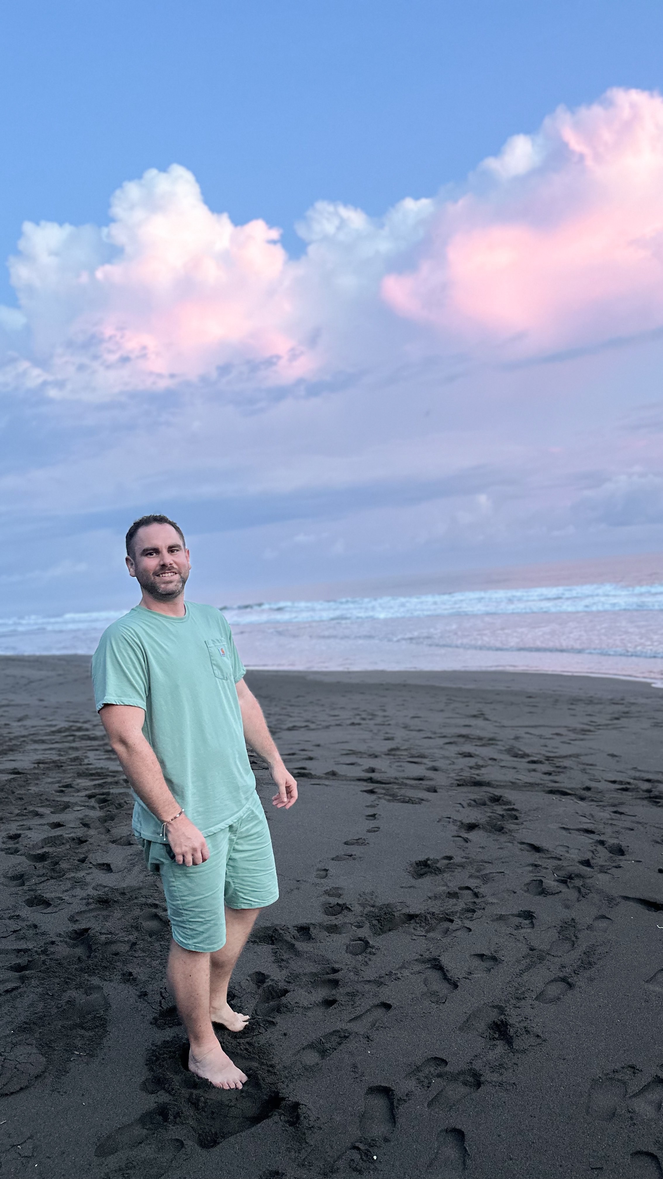 avid, founder of Velora Travel Studio, standing on a black sand beach during a pink sunset, wearing a casual green outfit