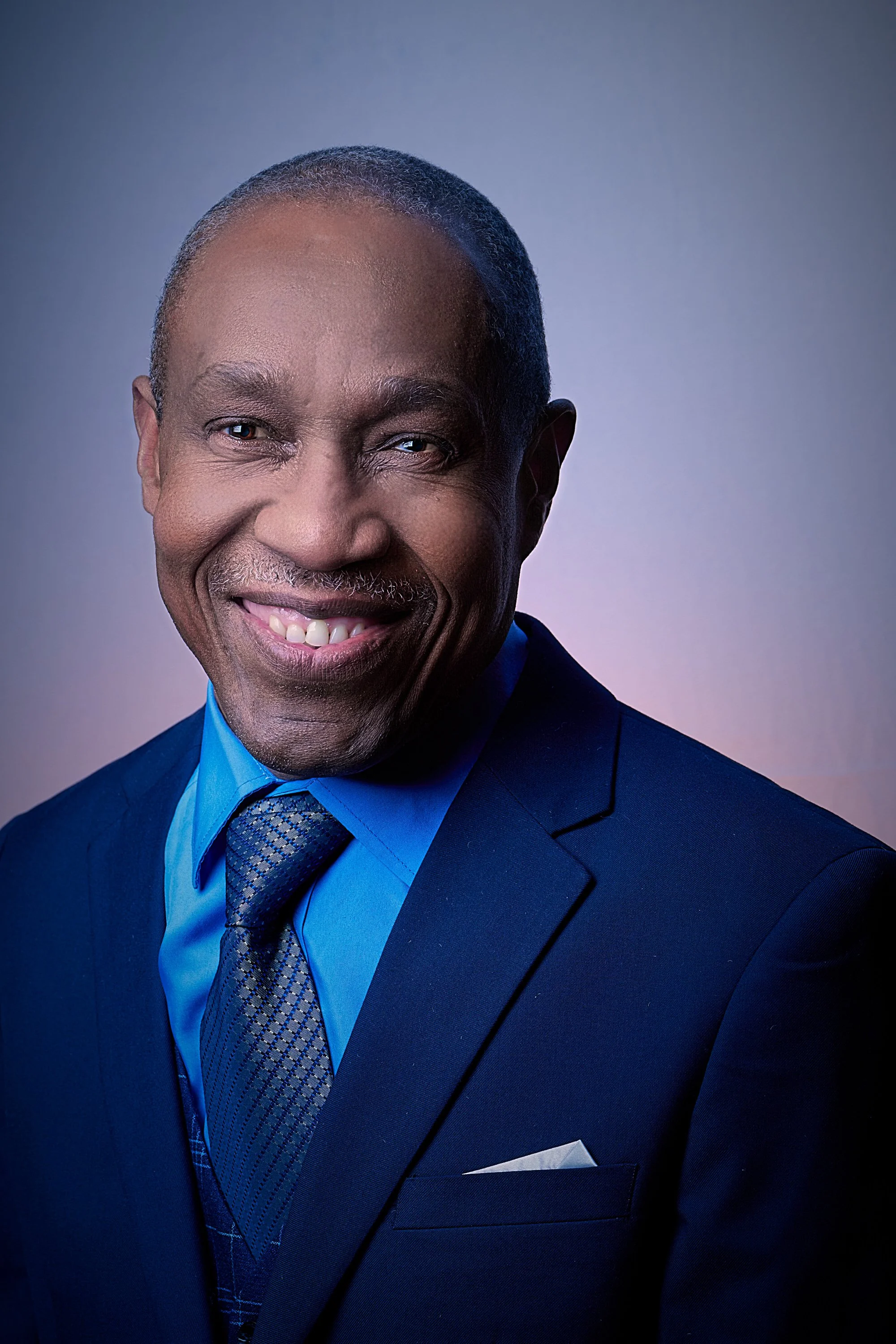A portrait of a smiling Black man wearing a dark suit, blue shirt, and patterned tie, standing against a gradient blue background.