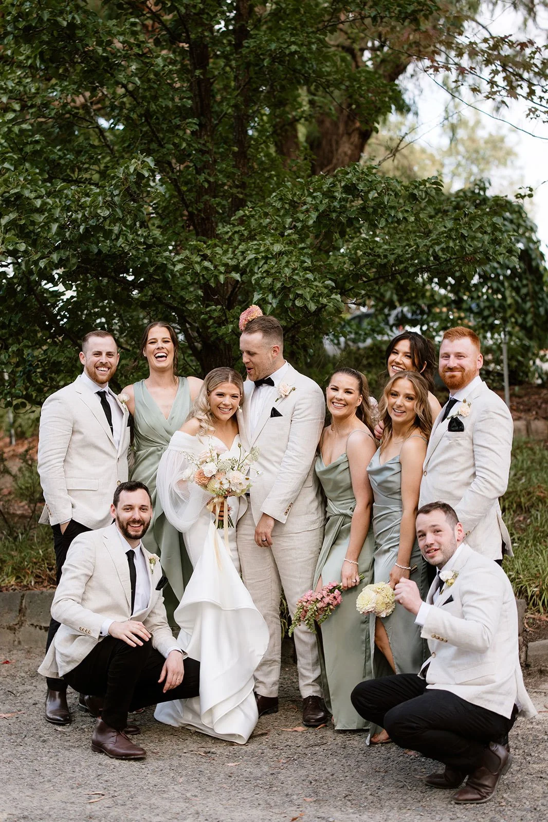 A group of ten people, including a bride and groom, standing outdoors in front of a large tree, dressed in wedding attire and posing happily for a wedding photo.