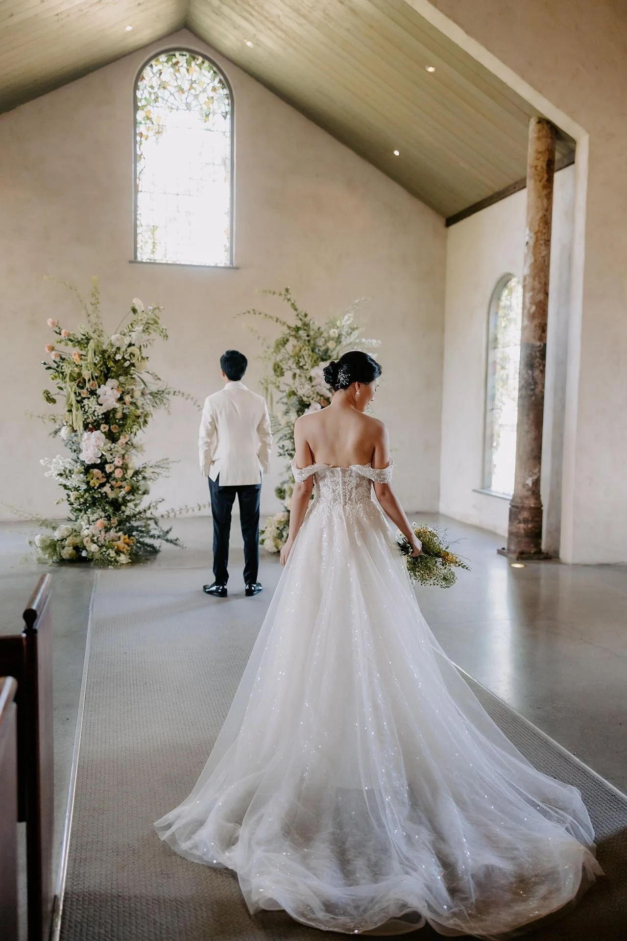 A bride in a white wedding gown holding a bouquet stands in a church. The groom is standing in front of her, facing away. The church has tall stained glass windows and floral arrangements.