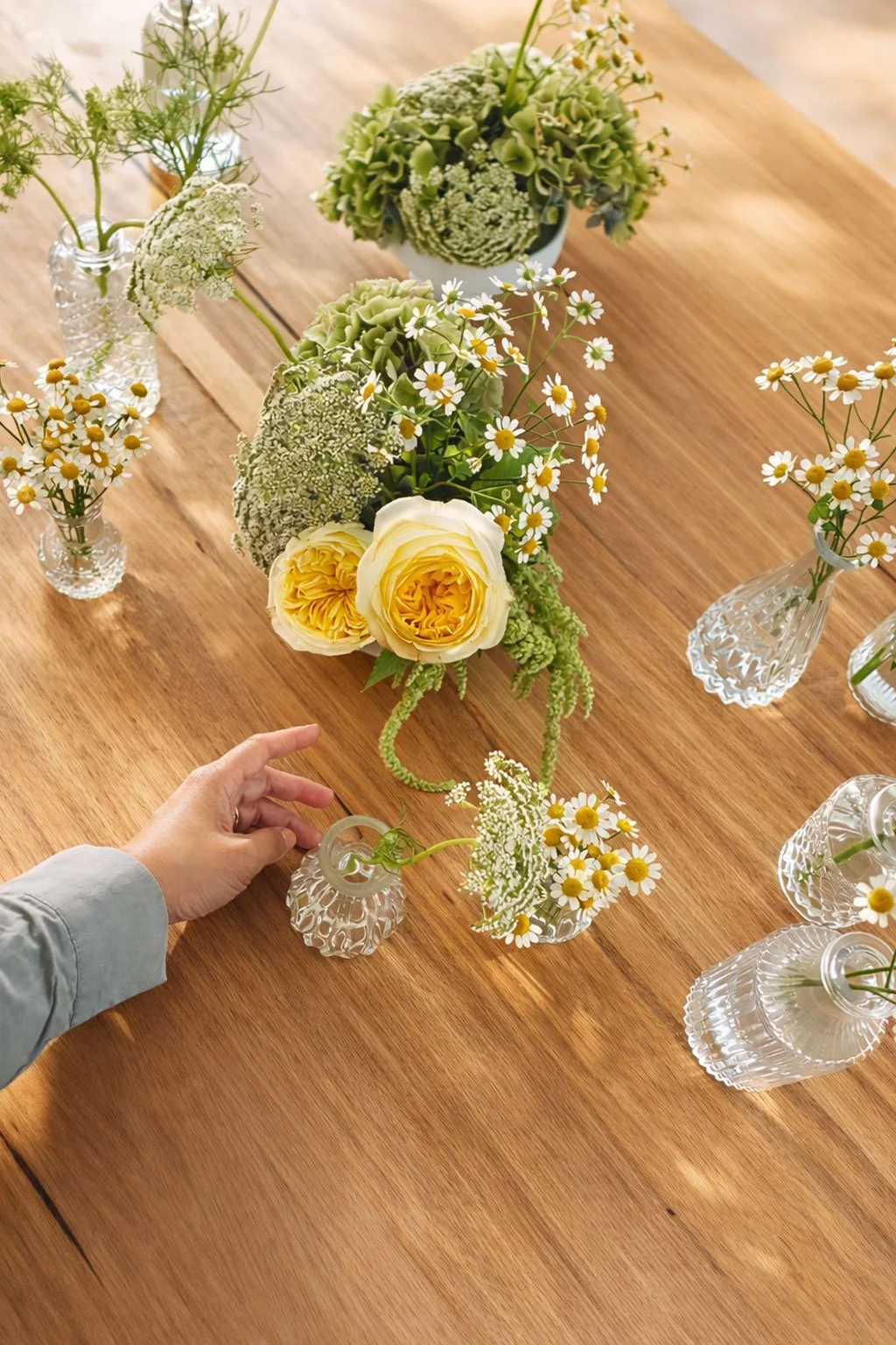 Person arranging small glass vases with white daisy-like flowers on a wooden table, with larger flower arrangements featuring yellow roses, white hydrangeas, and greenery.