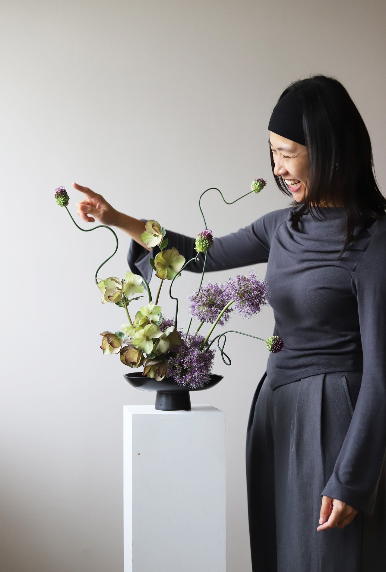 Workshop in Seoul: Leda in a grey outfit, smiling and pointing at a flower arrangement in a black vase on a white pedestal.