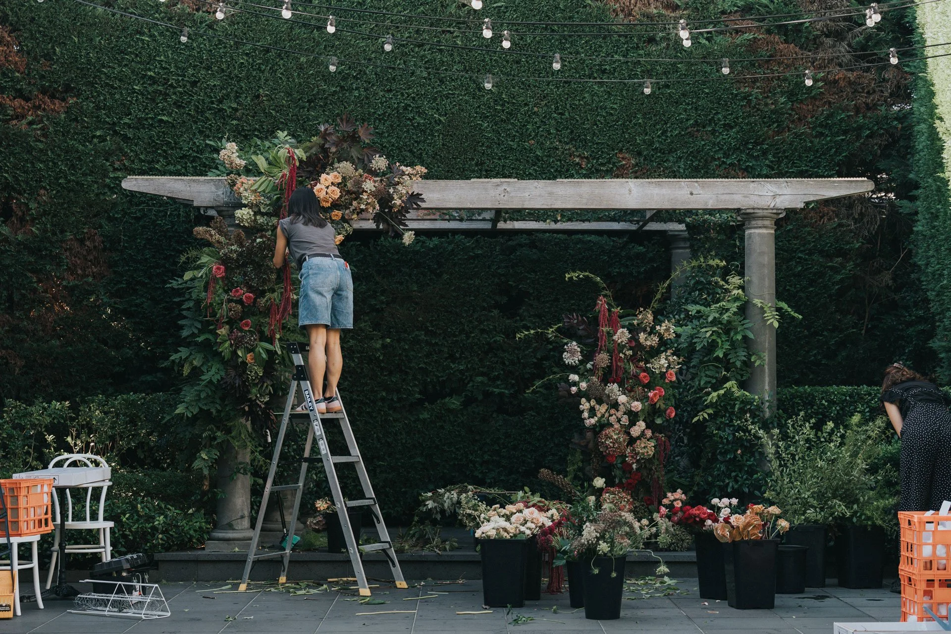 Two women arranging flowers on a floral arch in an outdoor garden setting with string lights overhead.