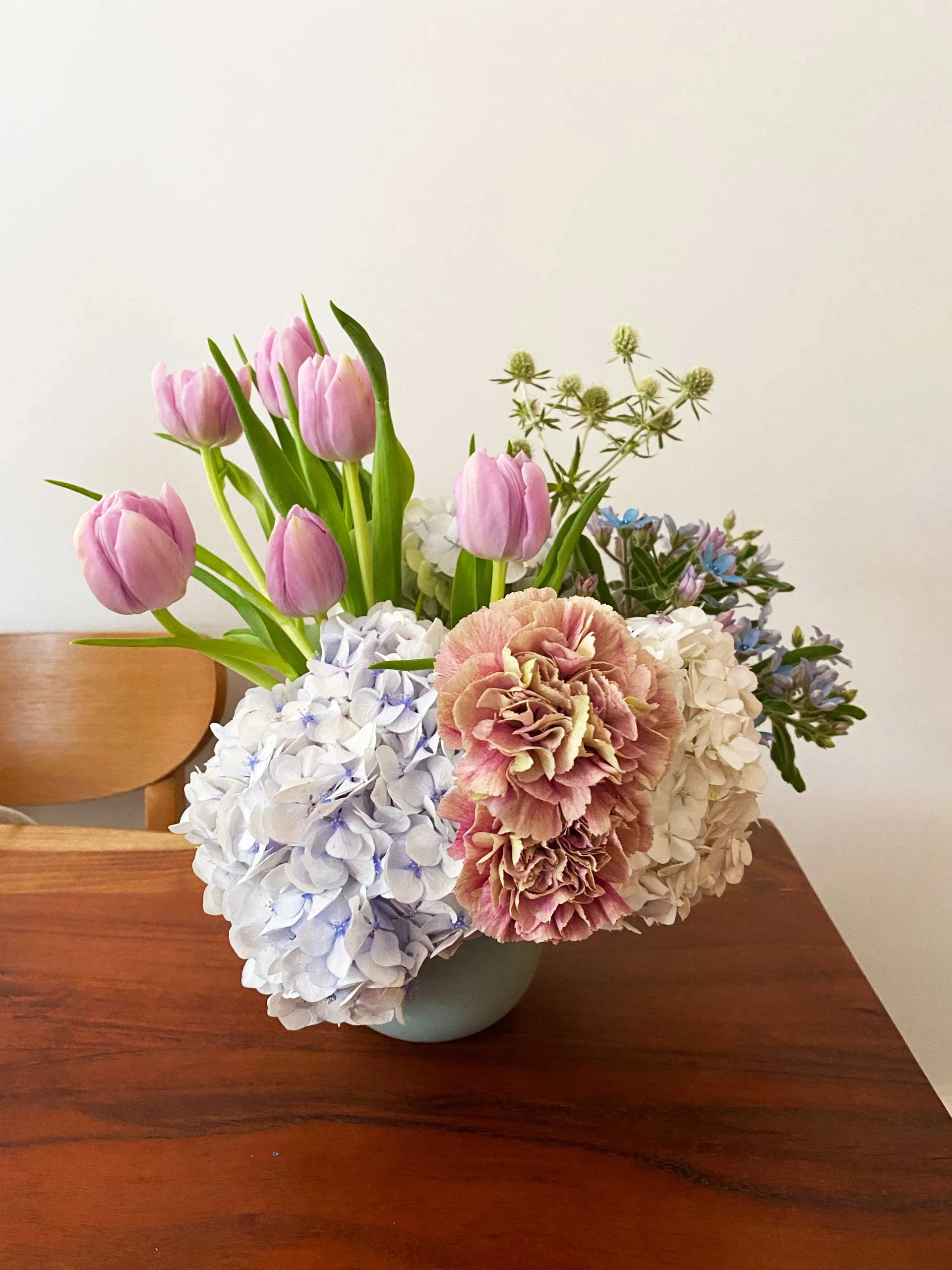 A bouquet of pink tulips, blue hydrangeas, and other assorted flowers in a light blue vase on a wooden table.