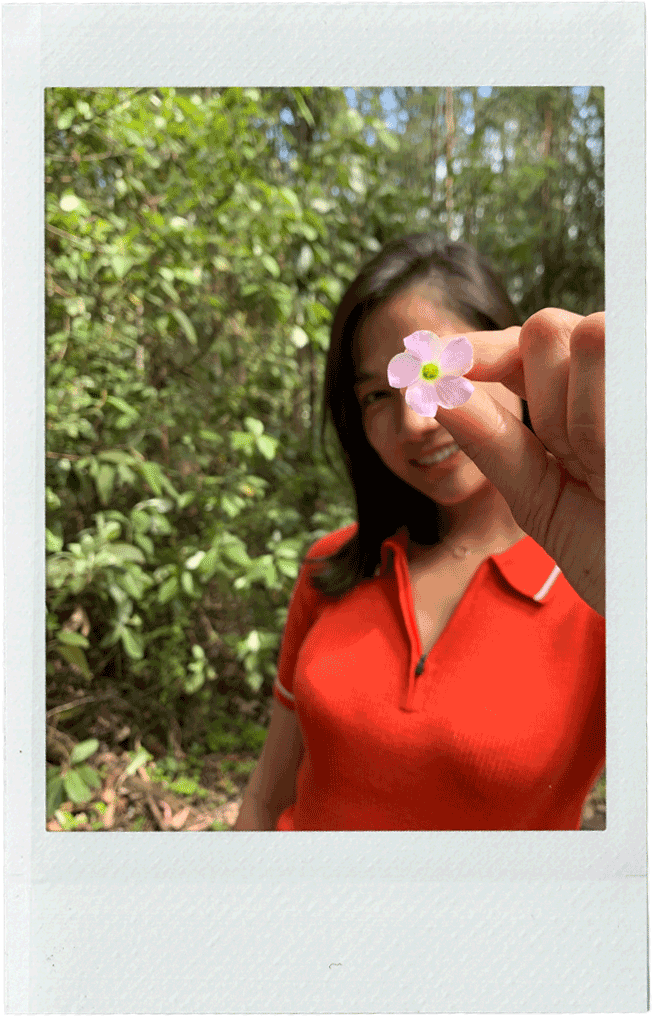 Leda in a red shirt holding a small pink flower in front of her face, with a greenery background.