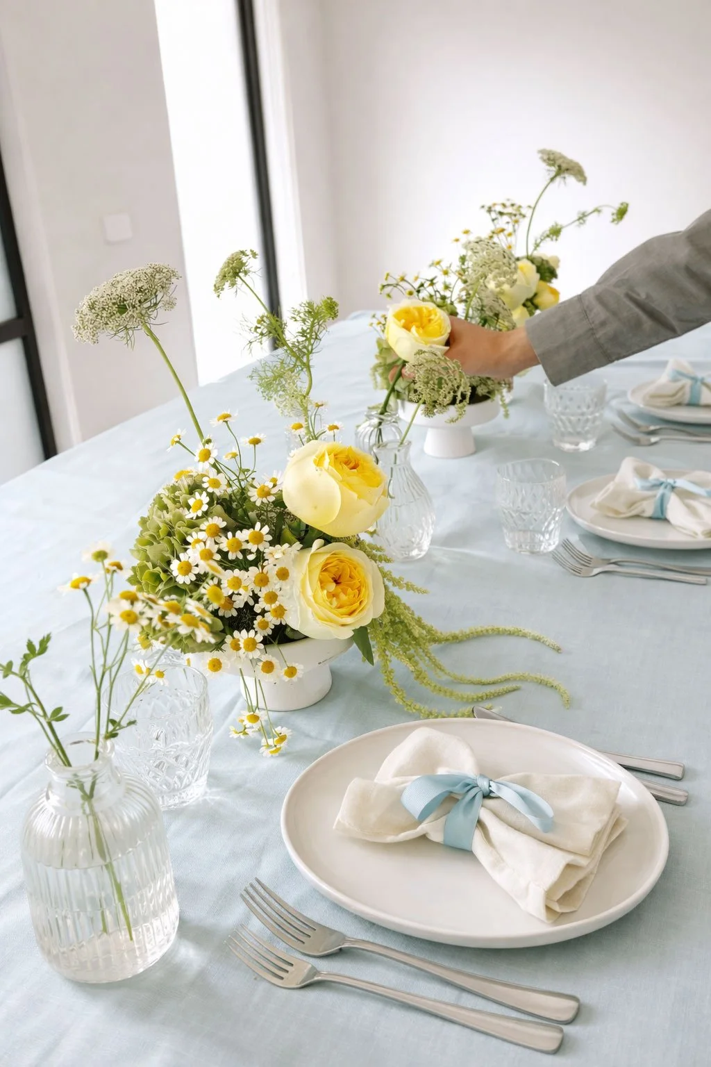 A table setting with vases of yellow roses, daisies, and greenery as centerpieces, with plates, napkins tied with light blue ribbons, forks, and glasses on a light-colored tablecloth.