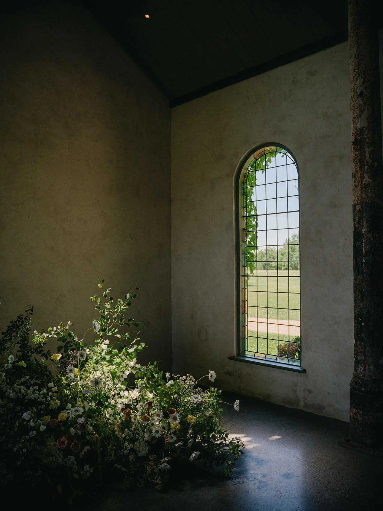 Interior view of a building with a tall, arched window, a flower arrangement on the floor, and sunlight streaming through the window, illuminating the room.