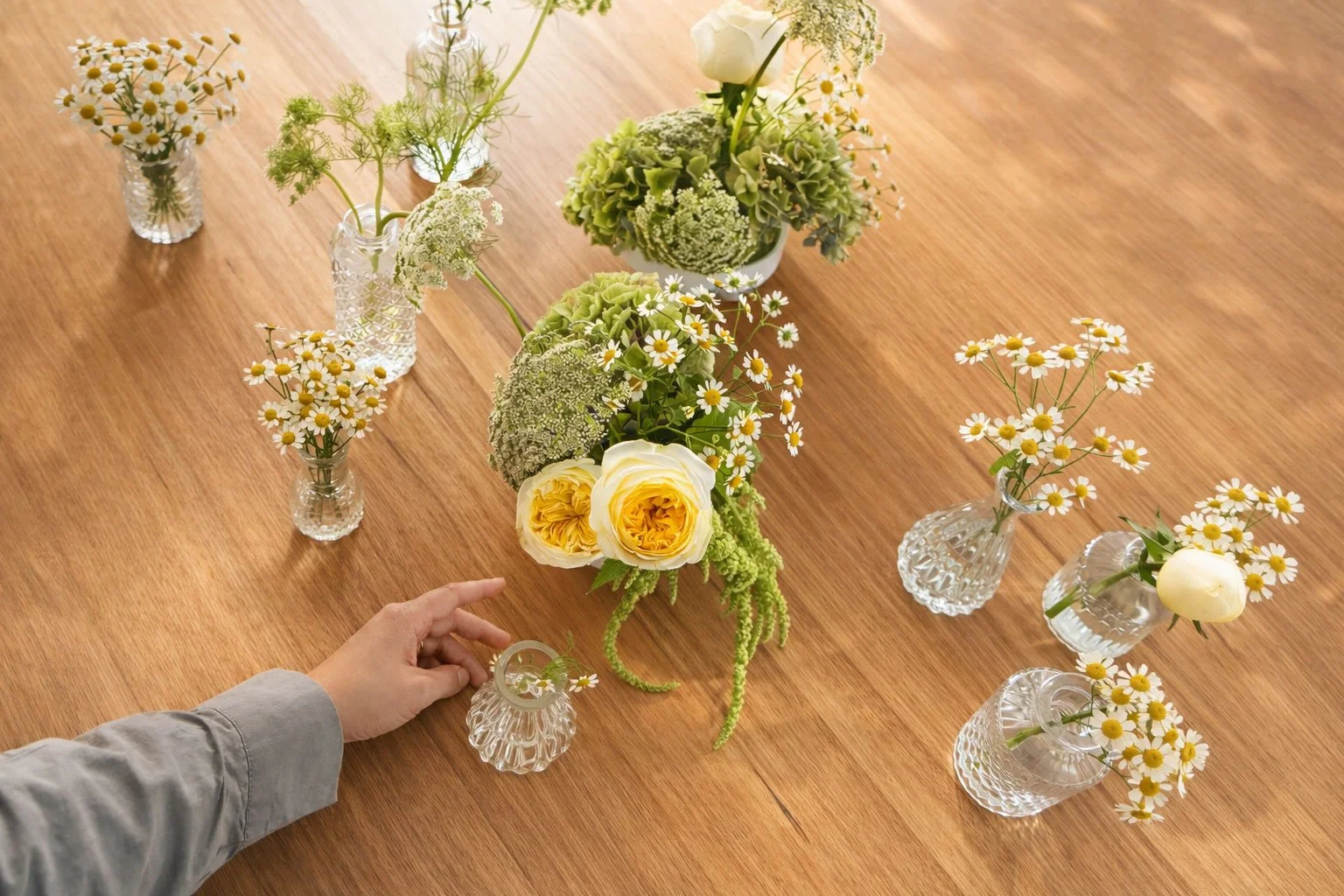 A person's hand adjusting a small floral arrangement in a decorative glass vase on a wooden table. The table has multiple small vases with white daisies, a larger vase with yellow roses, green hydrangeas, and other white flowers.