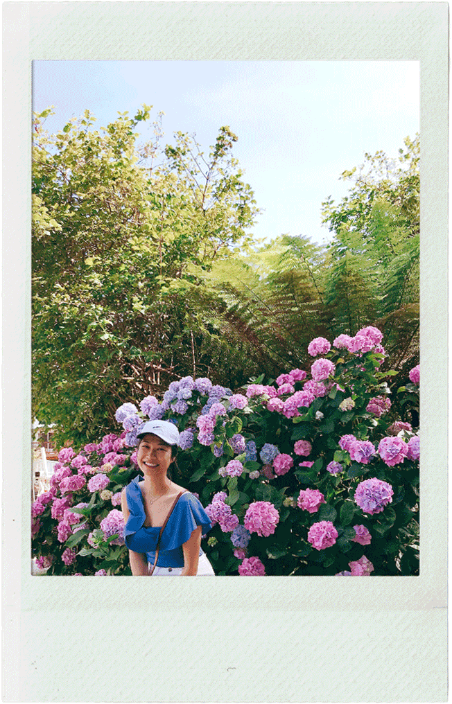 Leda with a white cap and blue dress smiling while standing in front of pink and purple hydrangea flowers in a garden on a sunny day.