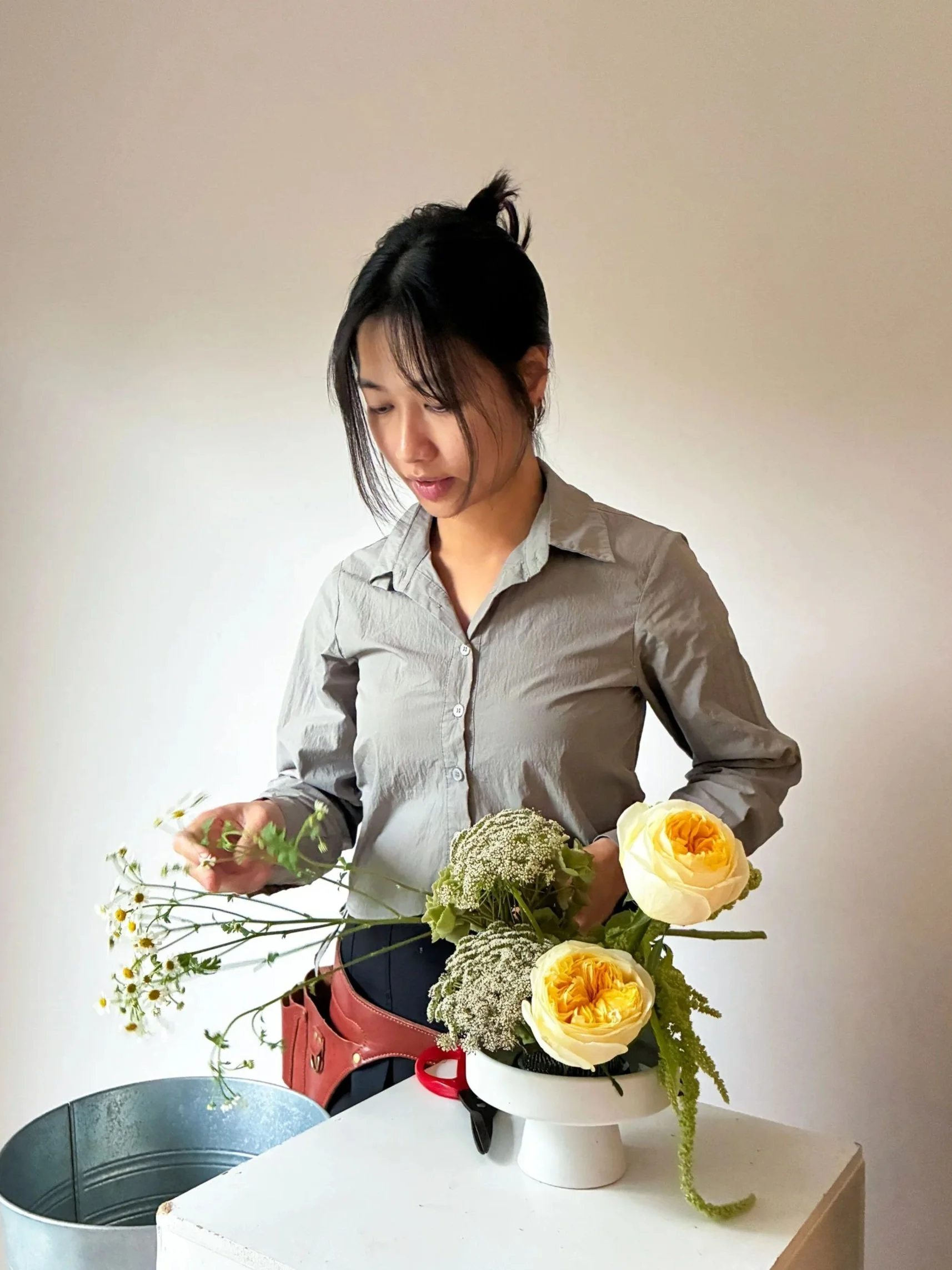 A woman arranging a flower bouquet with white and yellow roses and other greenery on a table.