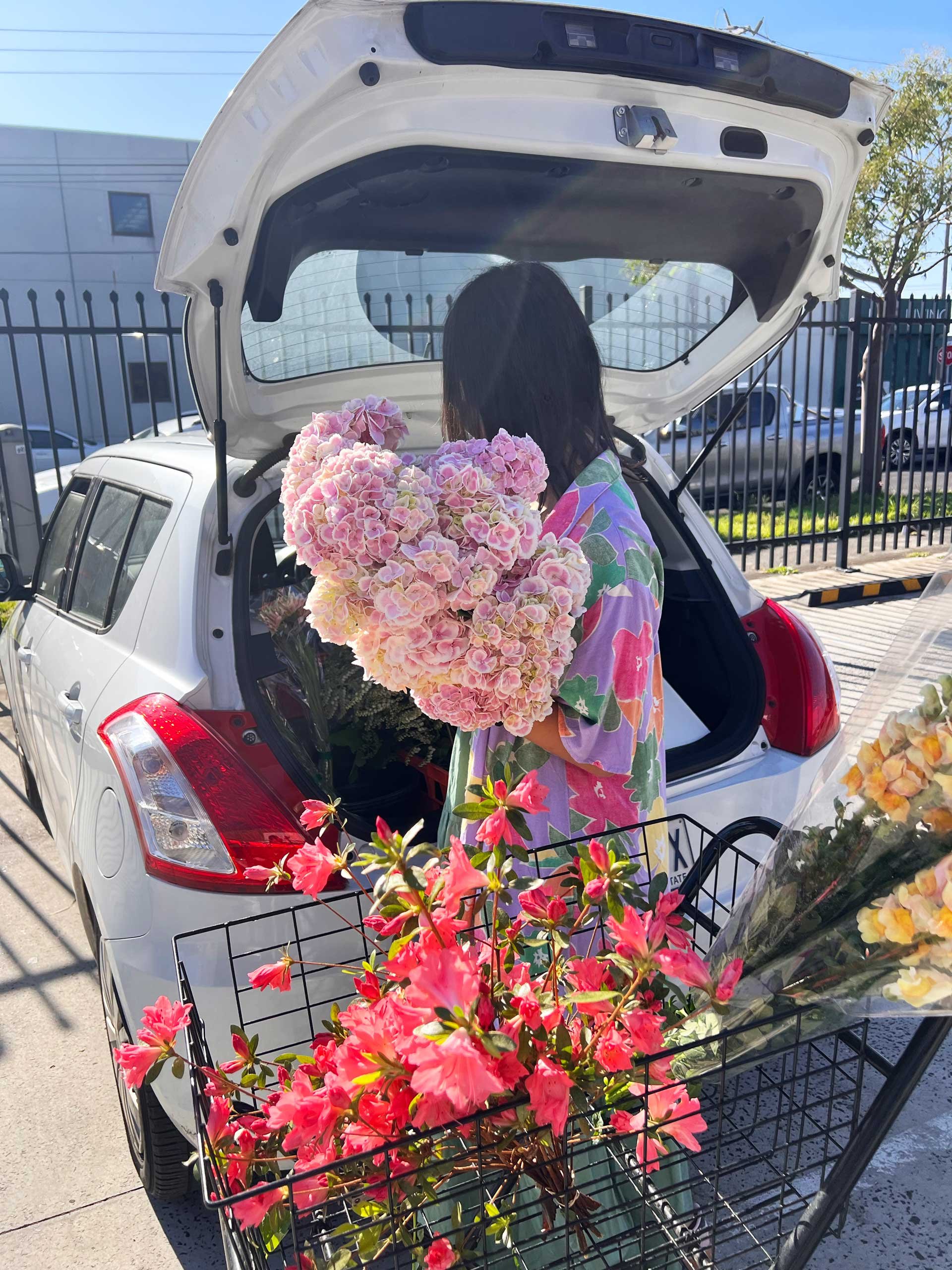 A person dressed in colorful floral clothing is standing in the open trunk of a white car, holding a large bouquet of pink and purple flowers. There is a shopping cart filled with bright pink flowers in front of the car, and a few more bouquets wrapped in clear plastic are visible. The scene is outdoors, with a metal fence, some buildings, and parked cars in the background.