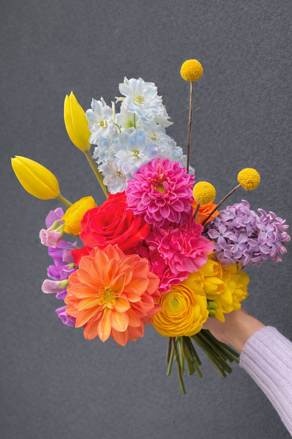 A hand holding a colorful bouquet of various flowers, including yellow tulips, white hyacinths, pink dahlias, purple lilacs, orange dahlias, and yellow ranunculus, against a gray background.