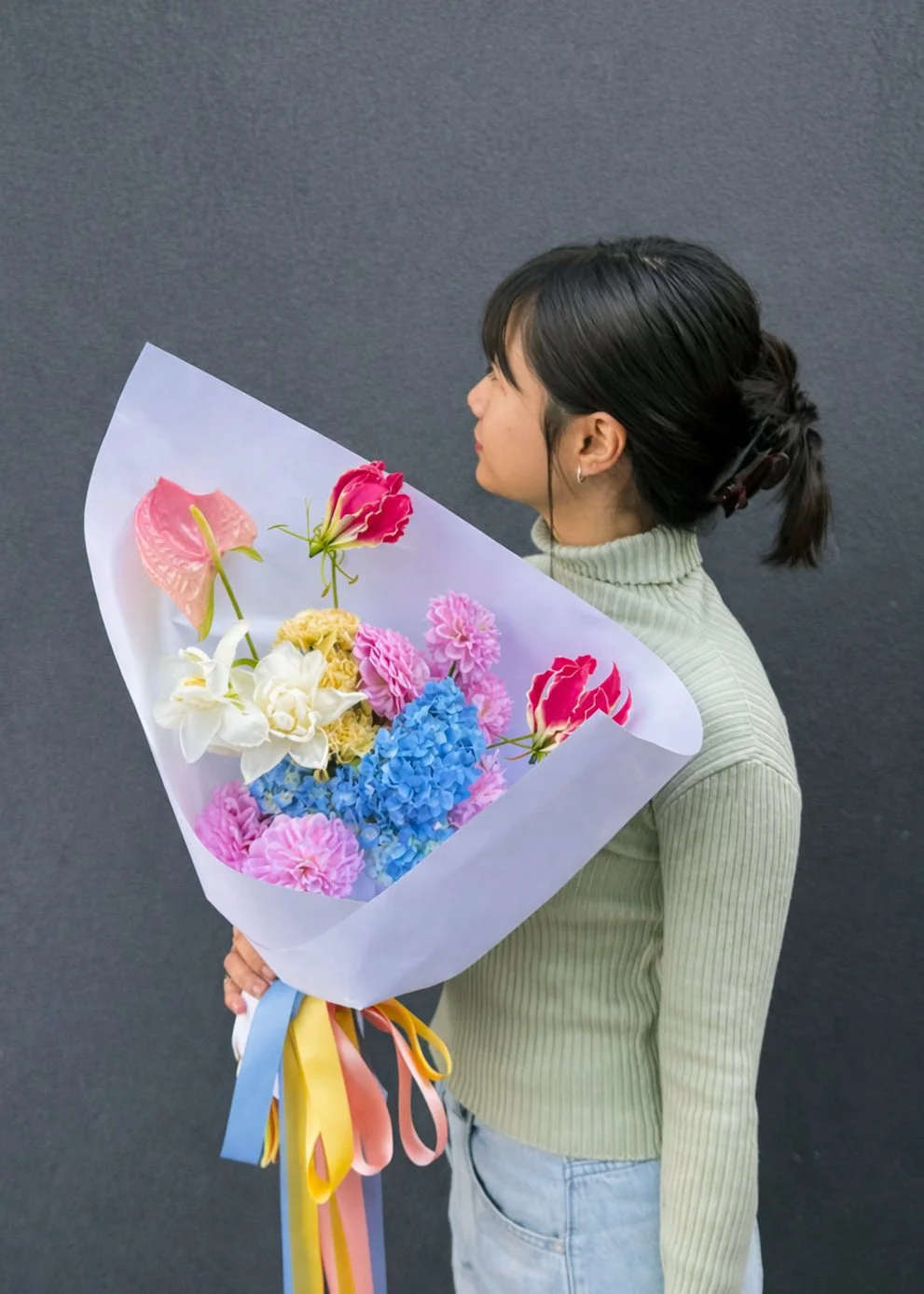 A woman in a green turtleneck sweater holding a bouquet of pink, blue, and white flowers against a dark gray background.