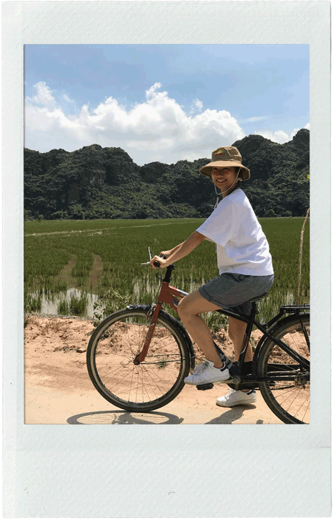 Biking in Northern Vietnam: Leda wearing a wide-brimmed hat, white t-shirt, grey shorts, and white sneakers rides a bicycle on a dirt path beside a green rice paddy field with mountains under a partly cloudy sky in the background.