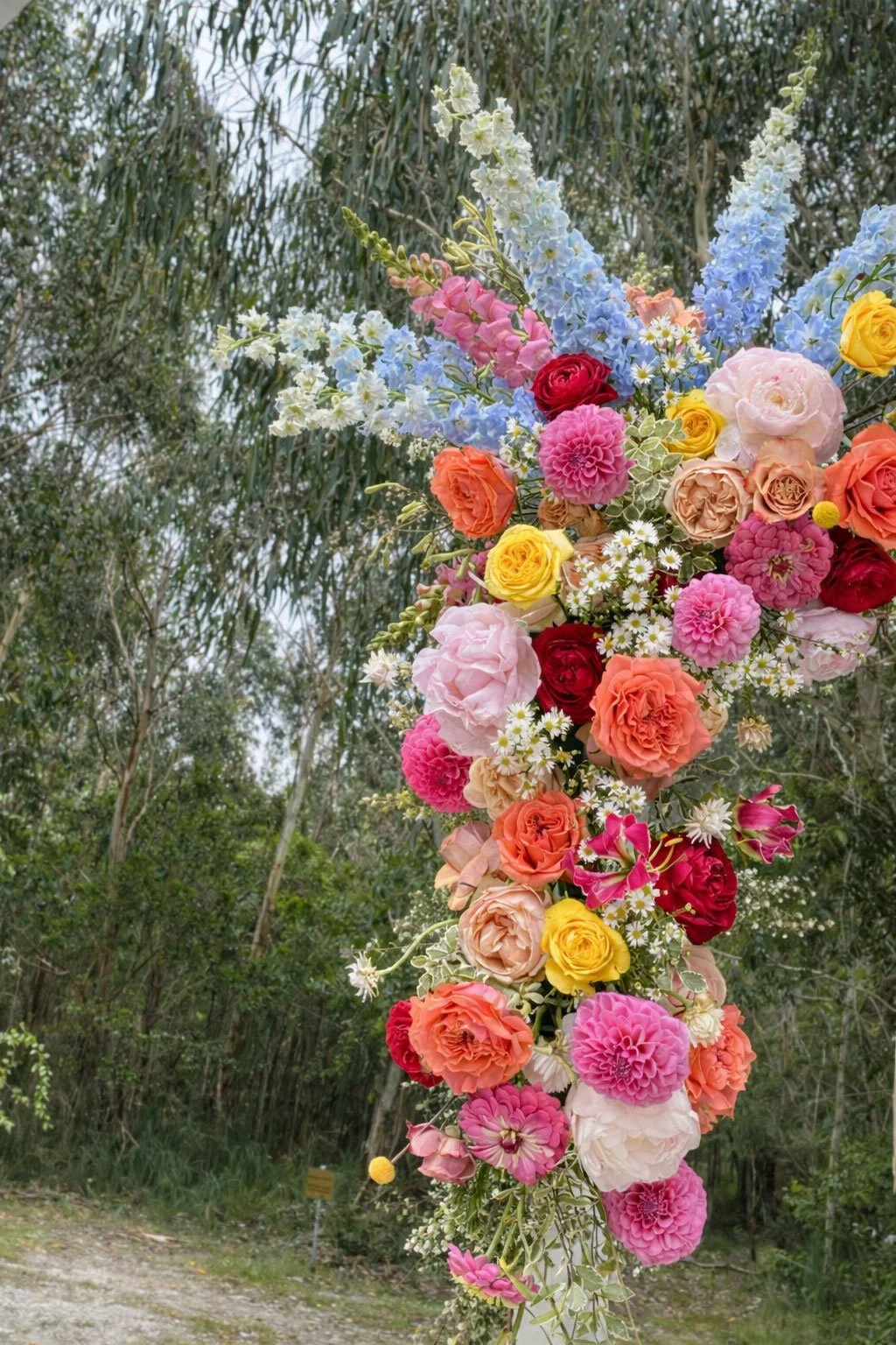 A tall floral arrangement featuring pink, red, yellow, peach, and blue flowers against a backdrop of trees and greenery.