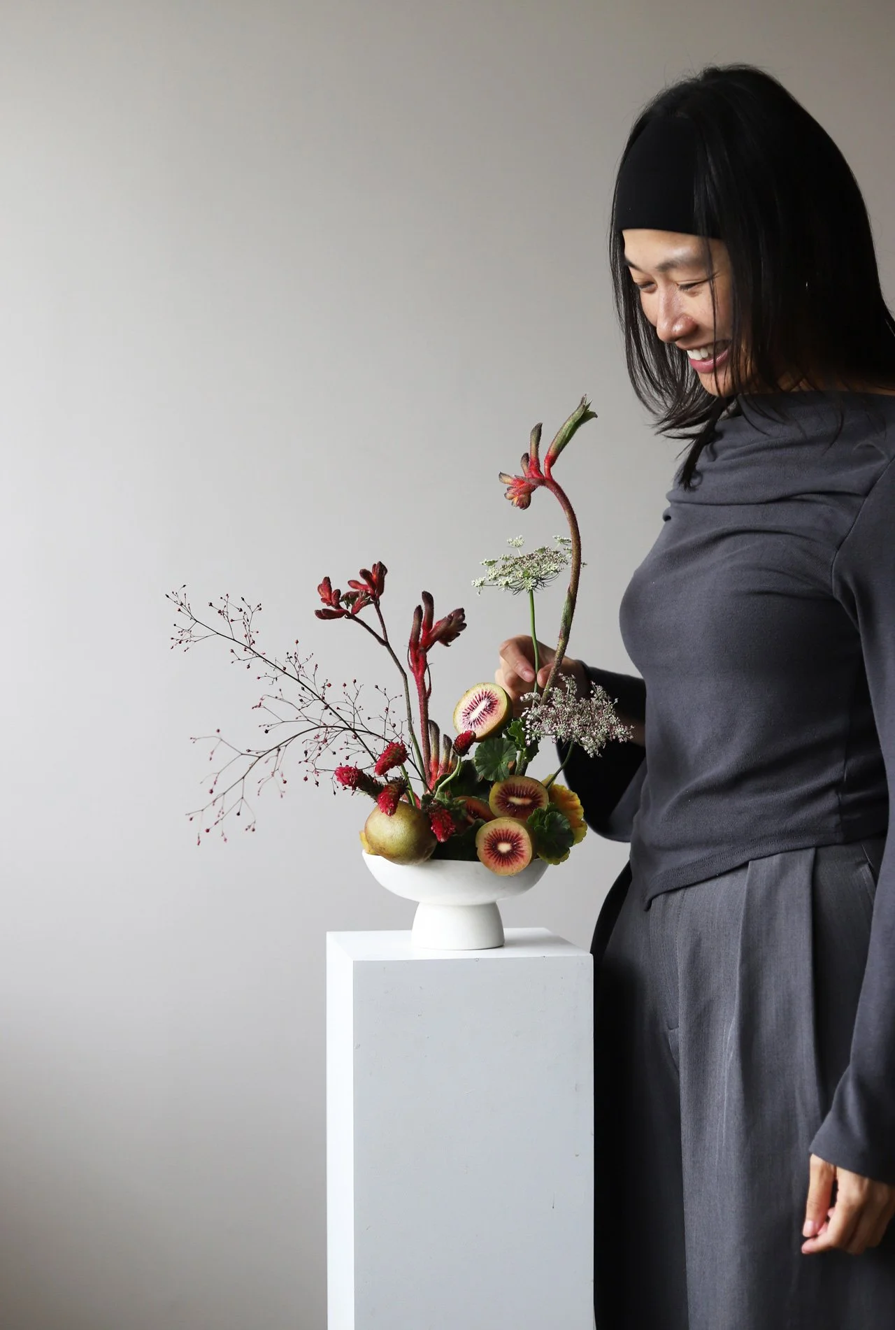 Workshop in Seoul: Leda in a grey outfit, is standing next to a white pedestal with a white vase containing a colourful floral arrangement including kiwifruit, red flowers, and various branches.
