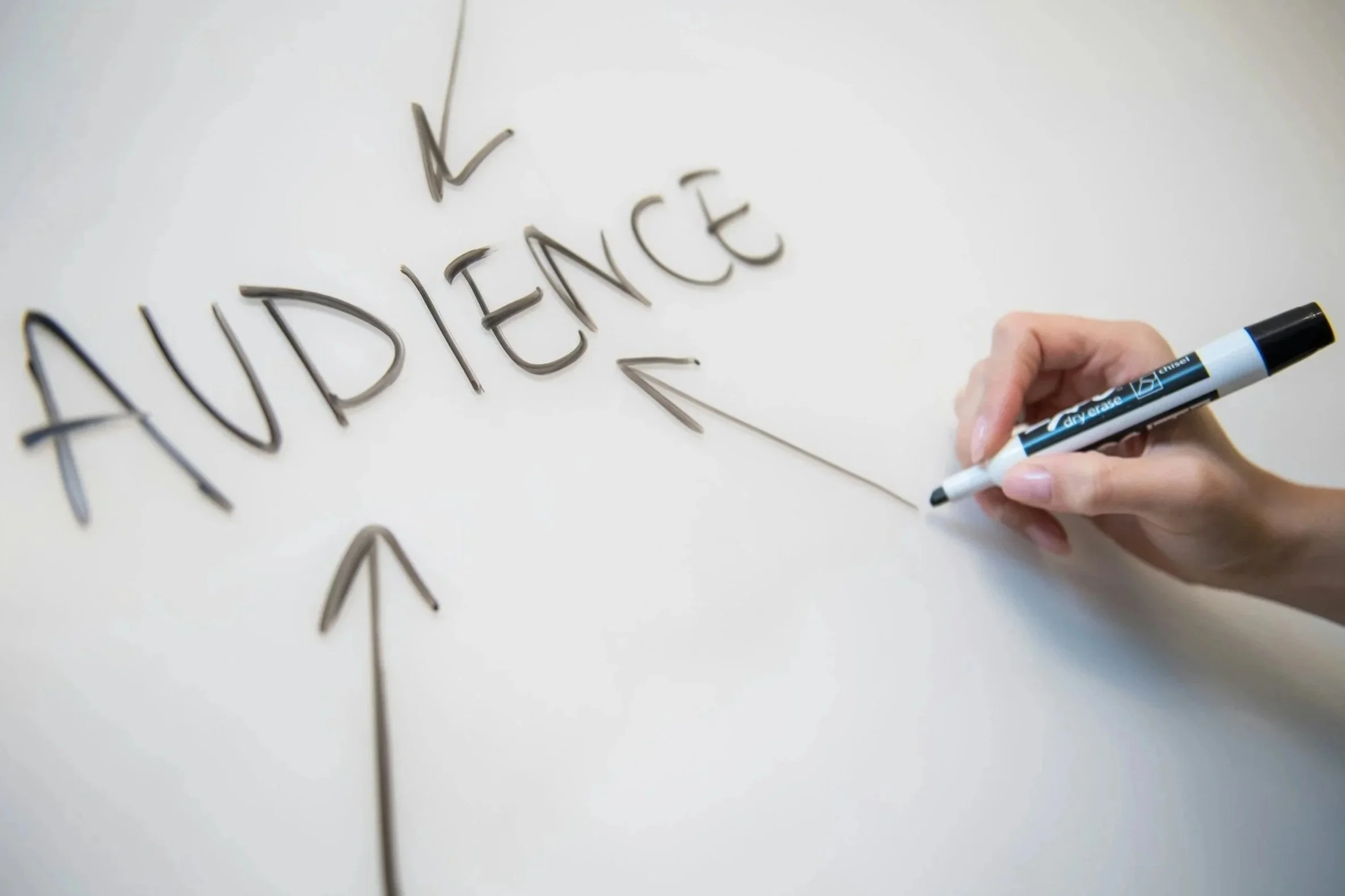 A hand writing the word 'AUDIENCE' on a whiteboard or paper with a black marker, with arrows pointing upward and downward.