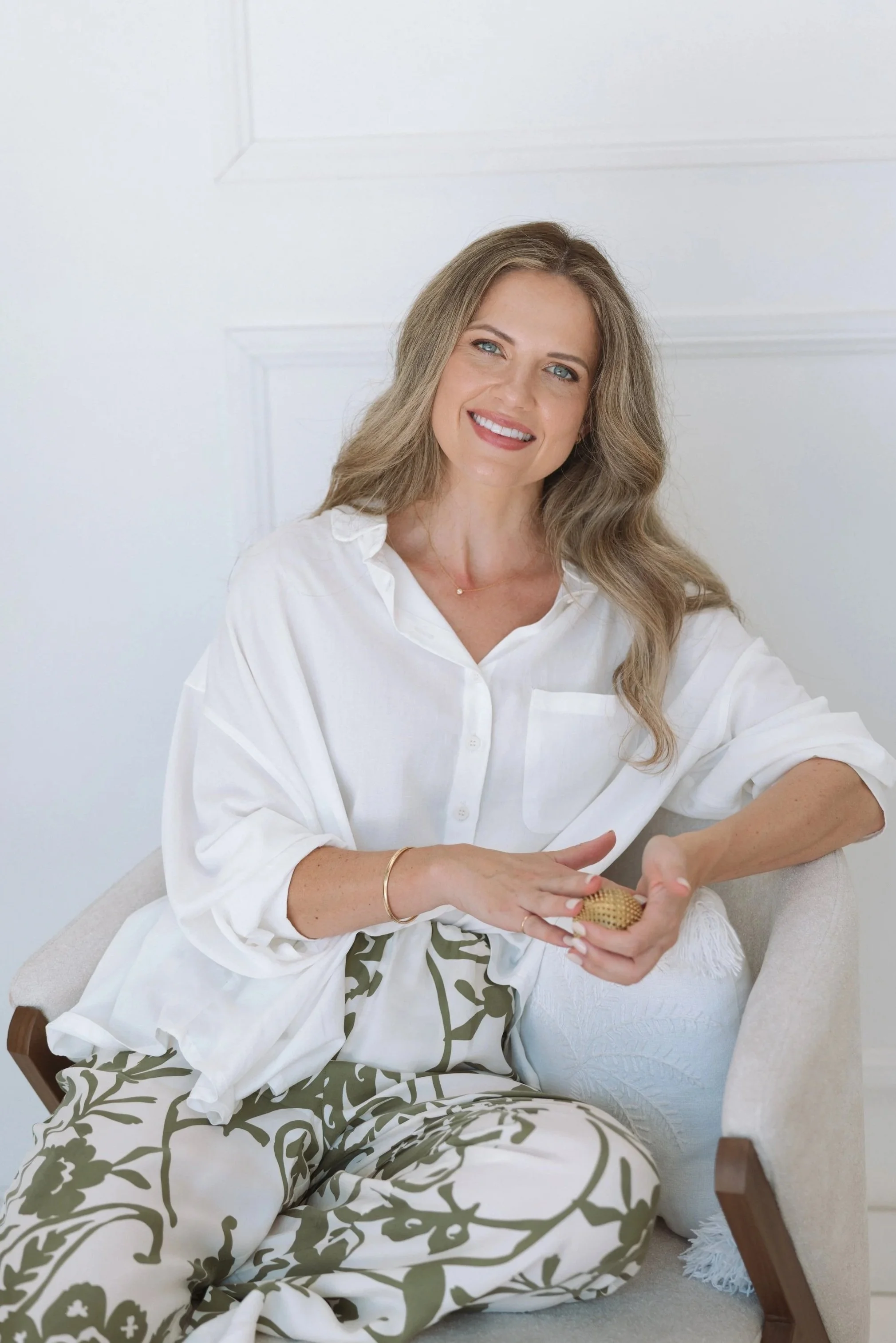 A woman with long, wavy blonde hair, wearing a white shirt and patterned pants, smiling and sitting on a white chair in a bright room with white walls.