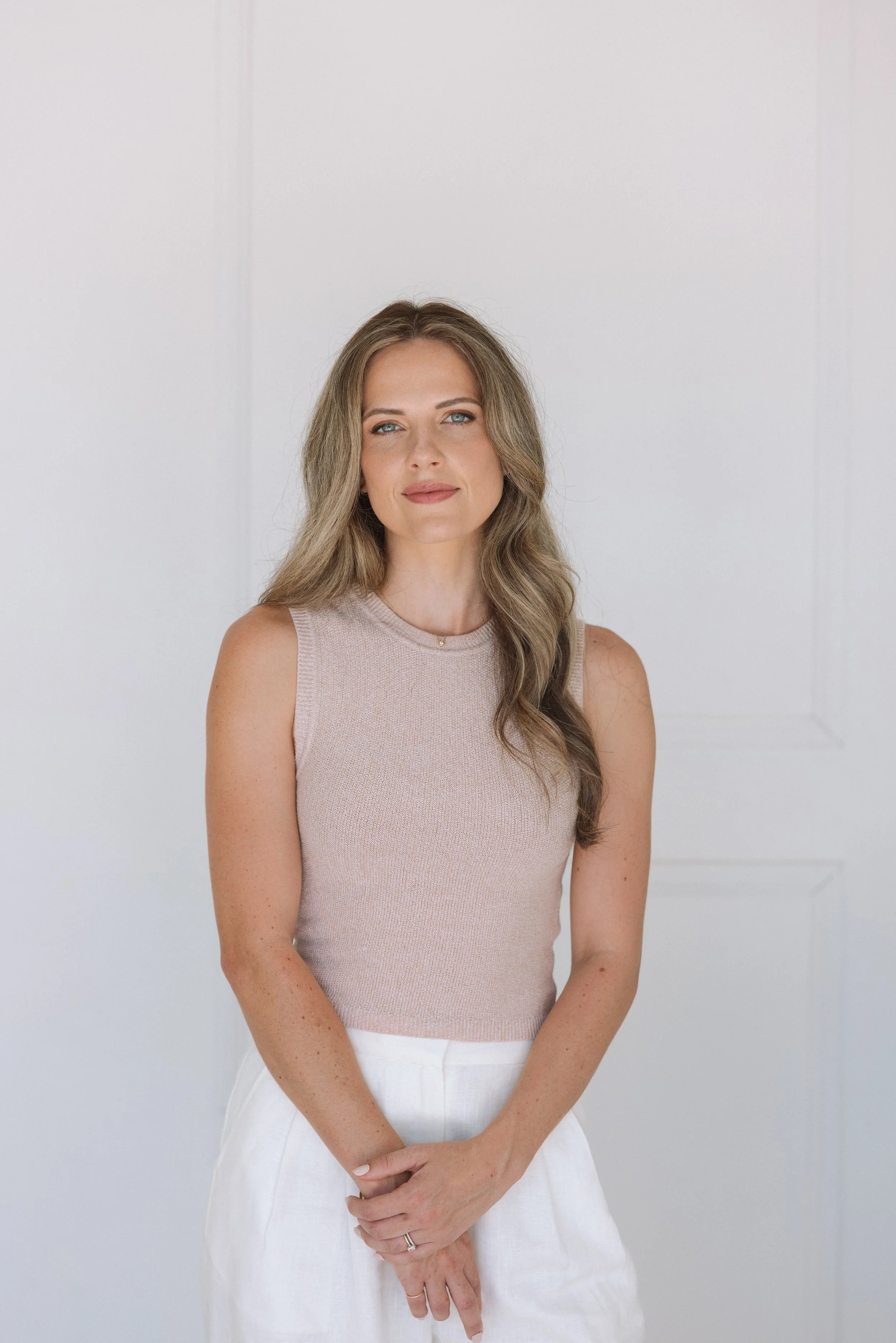A woman with long wavy hair and blue eyes wearing a sleeveless beige top and white pants, standing against a plain white background.
