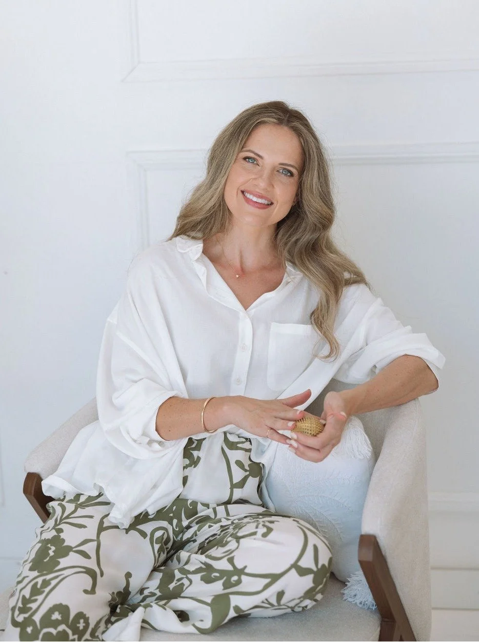 A smiling woman psychologist with long wavy blonde hair, wearing a white button-up shirt and patterned pants, sitting on a beige chair in a bright room.